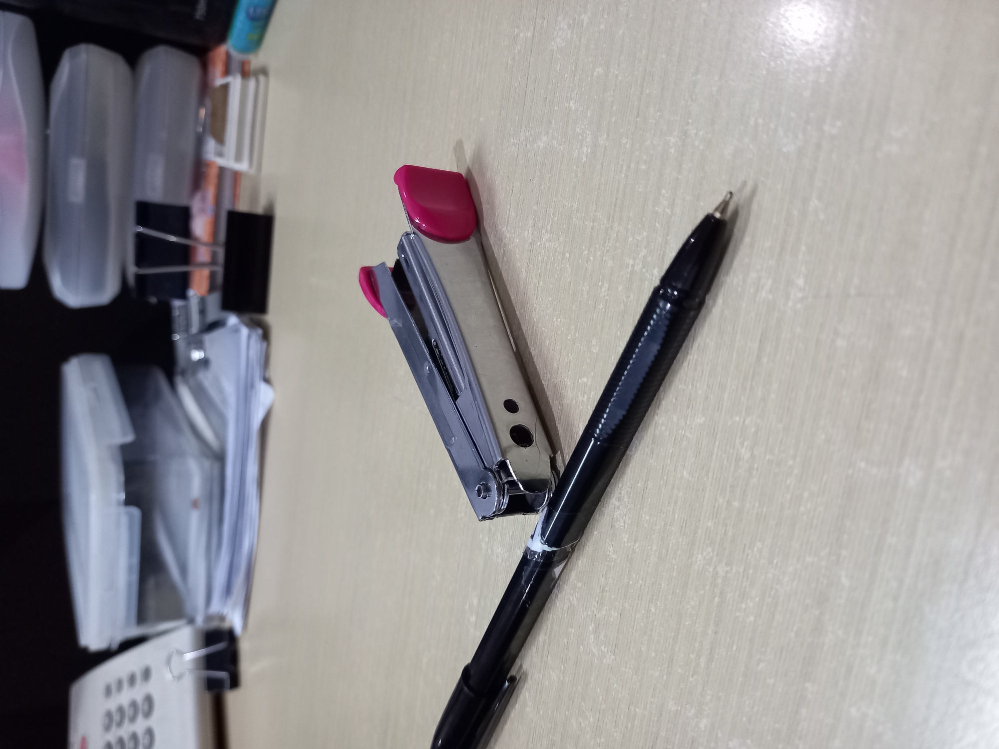 This is a close-up, eye-level, medium shot of a desk with office supplies. In the foreground, a black pen with a silver tip lies horizontally on a light-colored, wood-grain surface. To the right and slightly behind the pen is a silver stapler with pink accents. The stapler is positioned diagonally. In the background, to the left, a white telephone with gray buttons is visible, along with a black binder clip. Behind these items, there are stacks of papers and clear plastic containers. The lighting is dim, suggesting an indoor environment, possibly an office. The scene depicts a typical desk setup, without any indication of its specific location within Samarinda, Indonesia. There are no people or animals present.