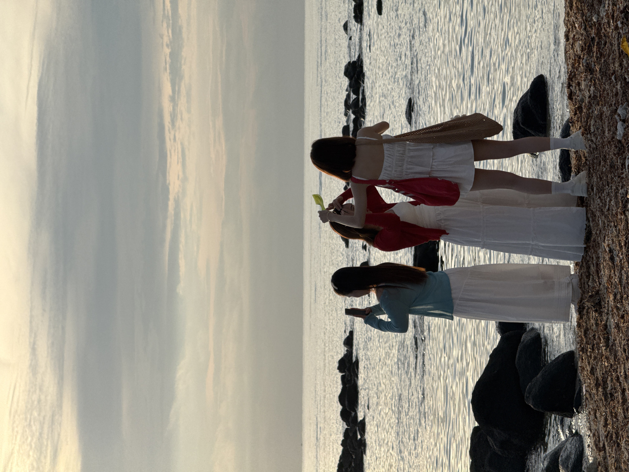Three individuals, appearing to be women, are standing on a rocky, sandy shoreline. The two individuals on the right are facing away from the camera, and the individual on the left is in profile, facing the sea. The person on the left is holding a mobile phone up, seemingly taking a photograph or video. The person in the middle is holding a bright yellow object above their head, possibly a fan or a prop for a photo, and appears to be interacting with the person on the right. The person on the far right is holding a woven bag on their shoulder and a red bag in their hand. All three are dressed in light-colored clothing, with two wearing white dresses and one wearing a blue cardigan over a white dress. The sea is calm with gentle ripples, and large, dark rocks are scattered along the shore and in the water. The sky above is overcast with soft clouds, suggesting either dawn or dusk. The location is the coast of Lý Sơn Island, Vietnam, a Special Administrative Region known for its volcanic landscapes and beaches.
