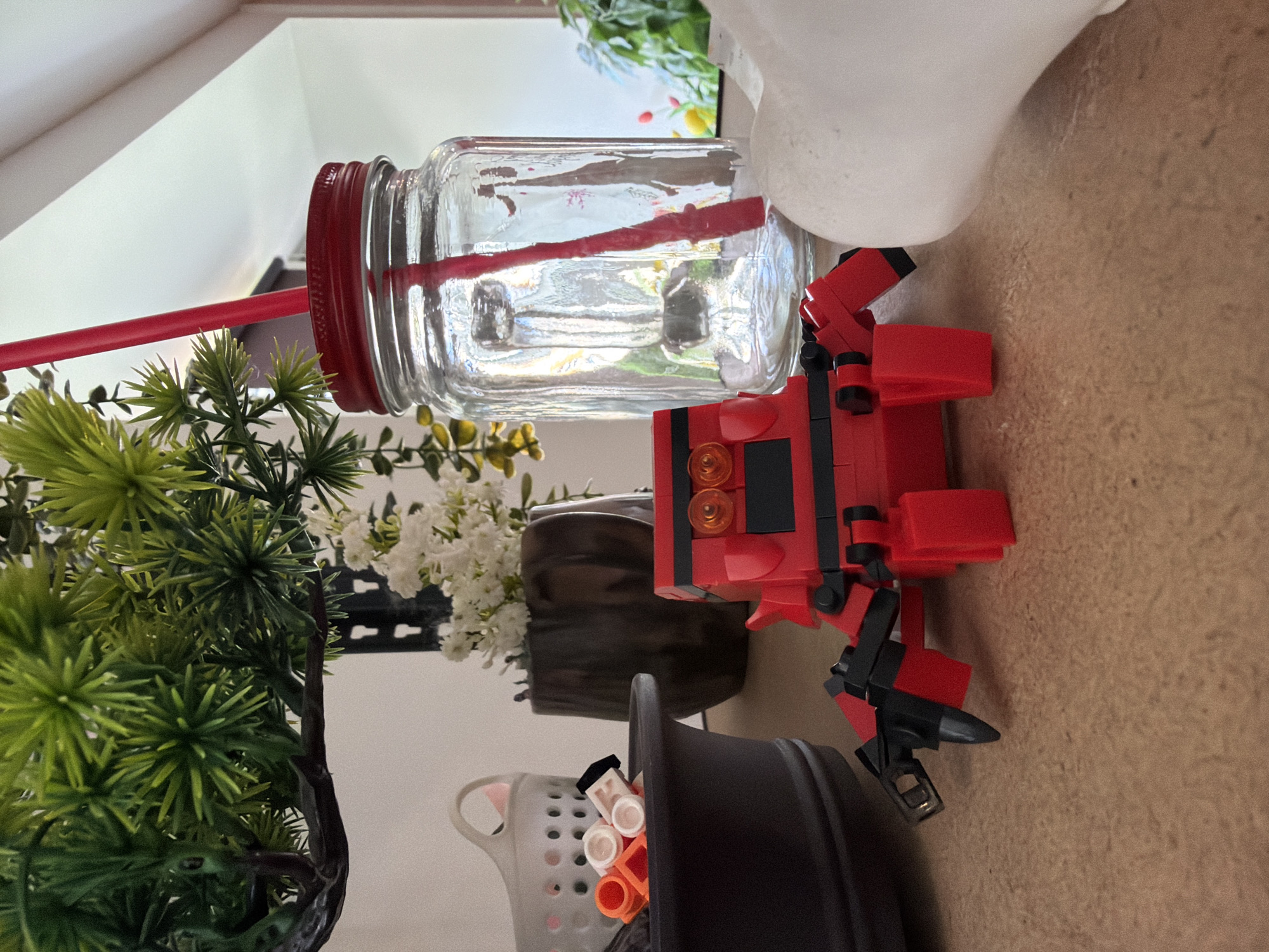 A close-up, low-angle shot focuses on a red LEGO robot toy positioned on a textured, light brown surface. The robot has black accents and appears to have robotic claws. To the right of the robot is a clear glass jar with a red lid and a red straw, partially filled with liquid. Behind the jar, a white, abstract object is visible. To the left of the robot, a dark gray pot contains a collection of small orange and white LEGO pieces. Further back, a dark brown pot holds a bouquet of small white flowers, and a faux green plant with needle-like leaves is situated behind that. The background is a plain white wall. The image does not depict any elements that would allow for identification of its location in Chaikovsky, Russia, or any specific city.