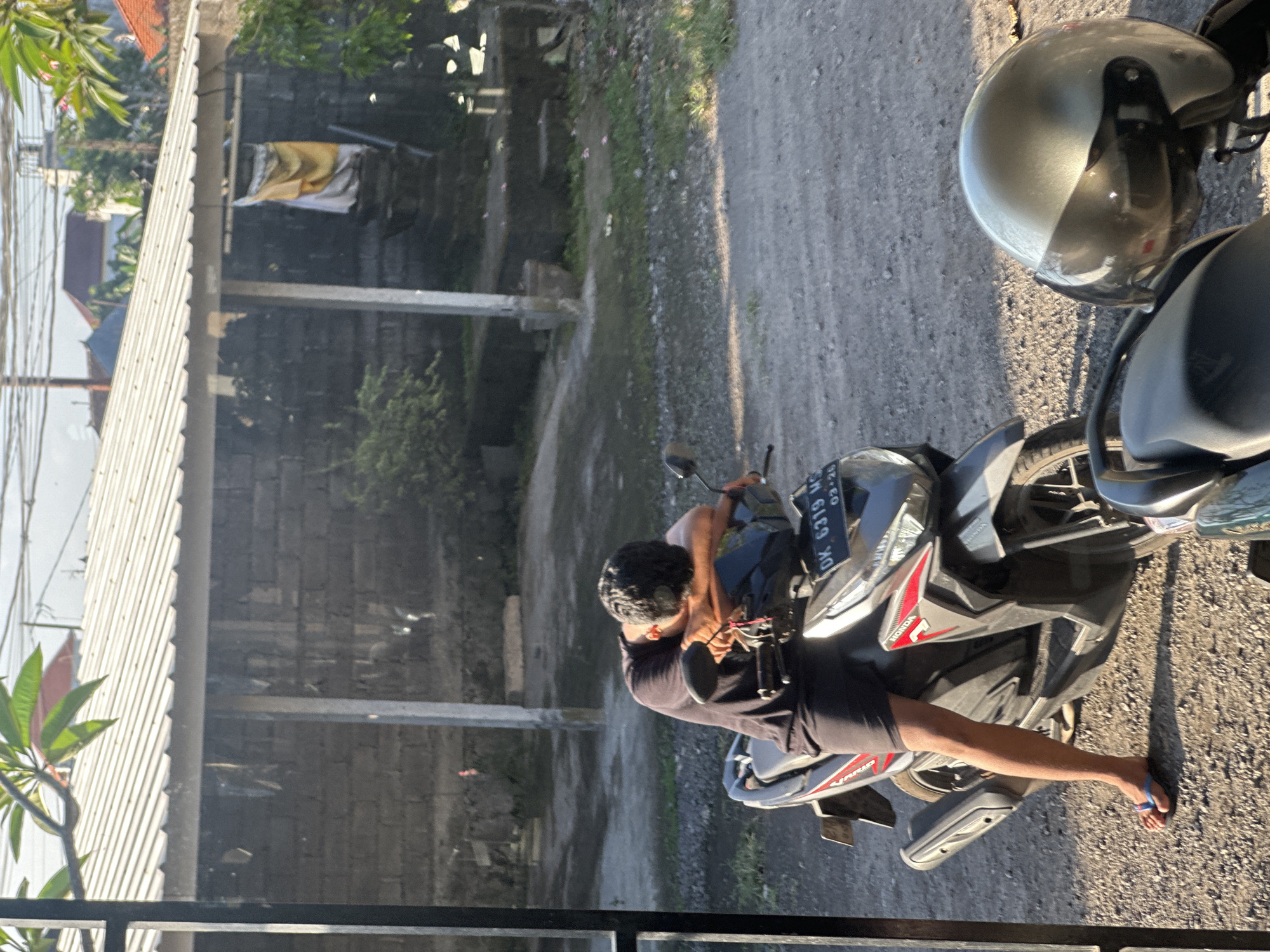 A man in dark shorts and a black t-shirt is asleep on a motorcycle, his head resting on his arms which are draped over the handlebars, in an outdoor area of Sukawati, Indonesia. The motorcycle, a Honda Vario, has a license plate reading "DK 6319 MC". A silver helmet is visible on the seat of another motorcycle in the foreground. The scene appears to be a residential area, with a brick building and a corrugated metal roof visible in the background. The ground is covered in gravel and dirt, and there is some greenery and vegetation around. The lighting suggests it is either early morning or late afternoon.