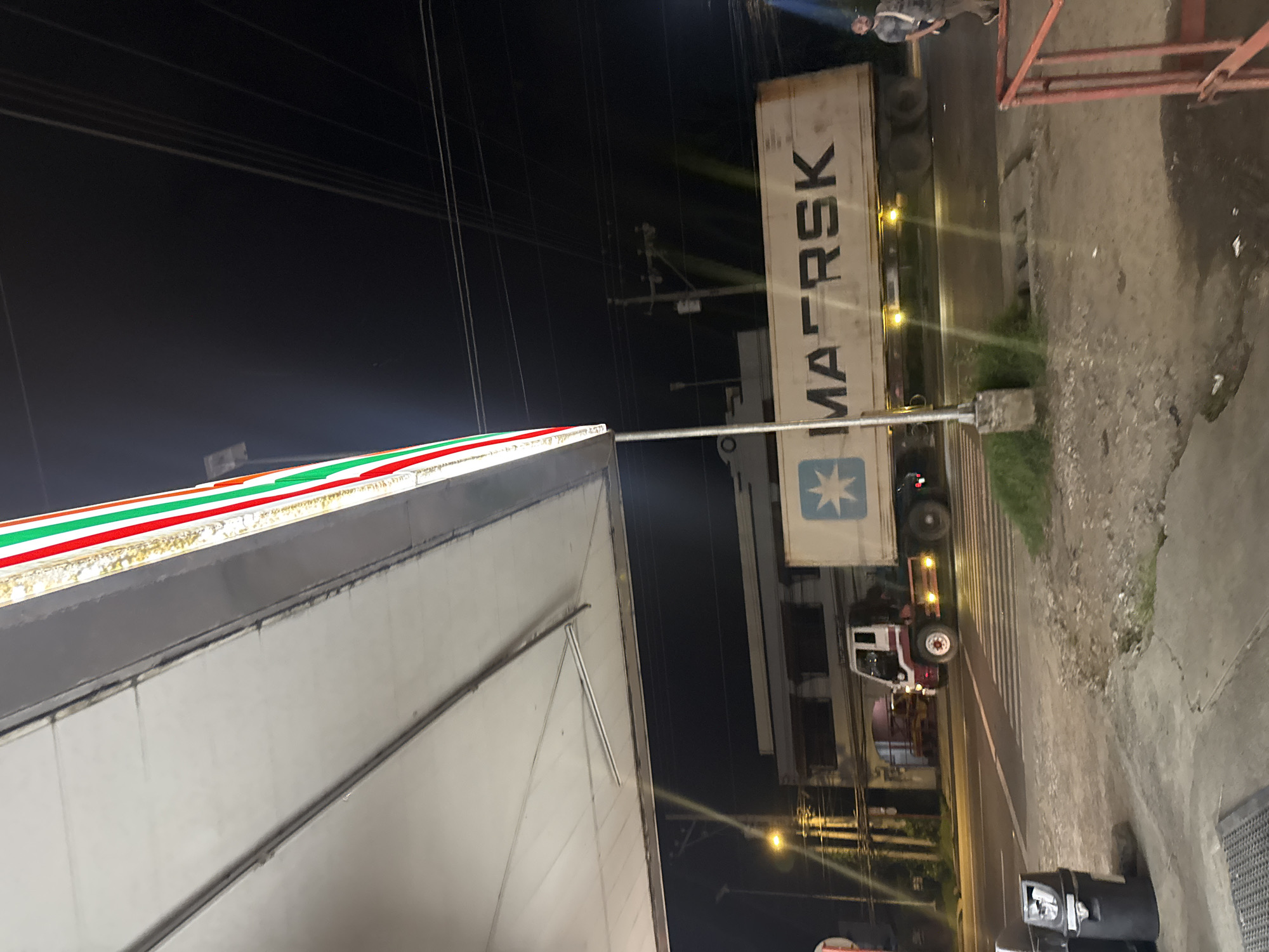 The media file captures a nighttime street scene in Davao City, Philippines, illuminated by artificial lights. The image is oriented vertically, showing a building on the left side with a distinctive green, white, and red striped facade, strongly resembling a 7-Eleven store. On the right, a large white shipping container labeled "MAERSK" in black, bold, vertically stacked letters is prominently displayed on the back of a truck. A white and red truck cab is partially visible below the container.
In the upper right portion of the image, a person wearing a light-colored top and dark pants is standing near the MAERSK container, looking towards the left. The scene includes a paved road with what appears to be a crosswalk or painted lines, and a patch of ground with some sparse vegetation near the truck. Utility poles and overhead power lines are visible against the dark sky, particularly in the upper and middle sections of the left side. Streetlights and vehicle lights cast reflections on the wet or damp road surface, adding to the nighttime ambiance. A metal pole extends upwards from the ground near the center of the frame, possibly part of a signpost or street light. The overall atmosphere is urban and industrious.