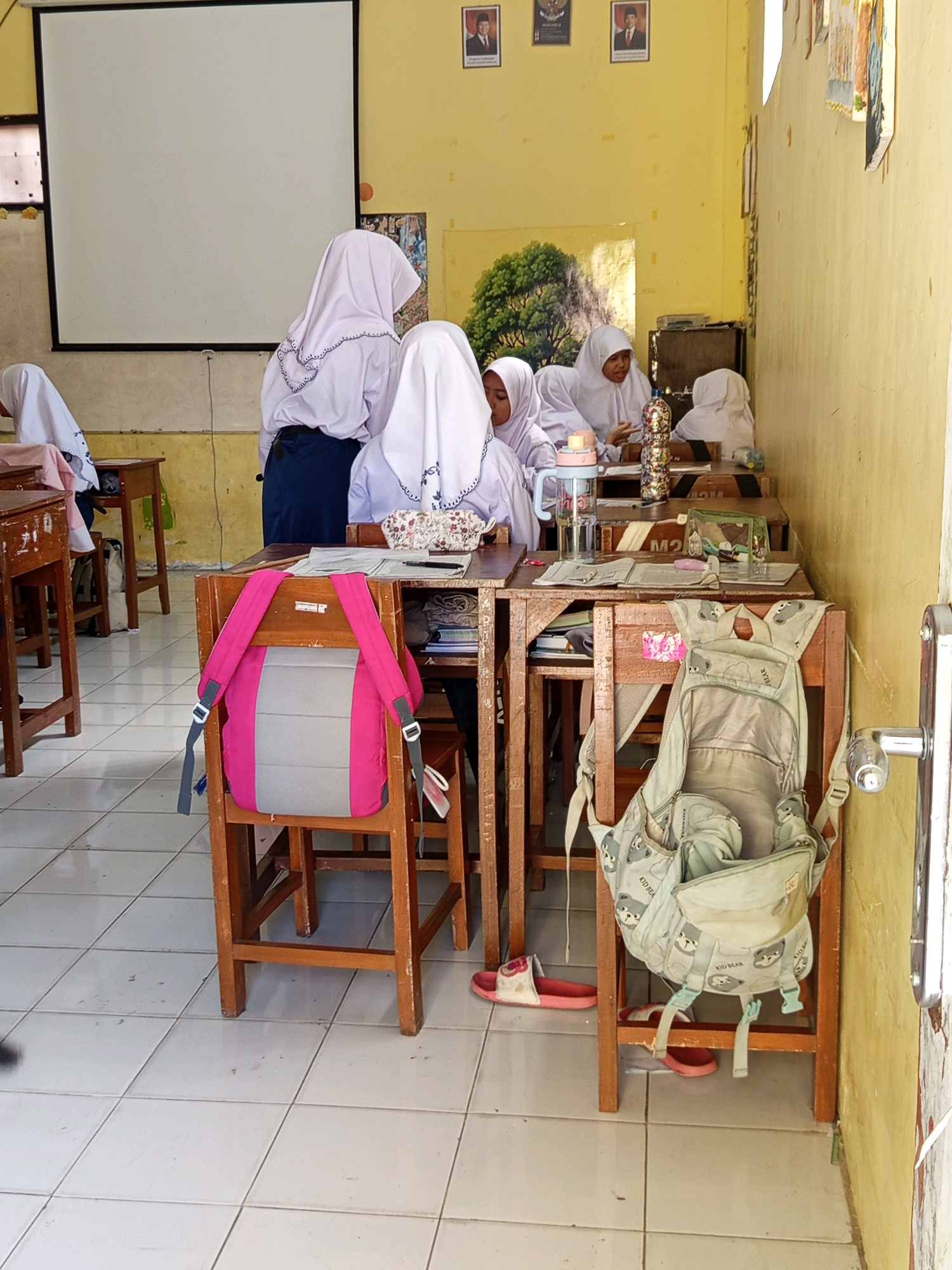 A classroom in Malang, Indonesia, is depicted with several students wearing hijabs and uniforms seated at wooden desks. A whiteboard is visible on the left wall, along with framed pictures of men in formal attire. The walls are painted a pale yellow. Backpacks, a water bottle, and a pencil case are present on the desks and chairs. A door handle is visible on the right side of the image. The floor is tiled with a light-colored pattern.