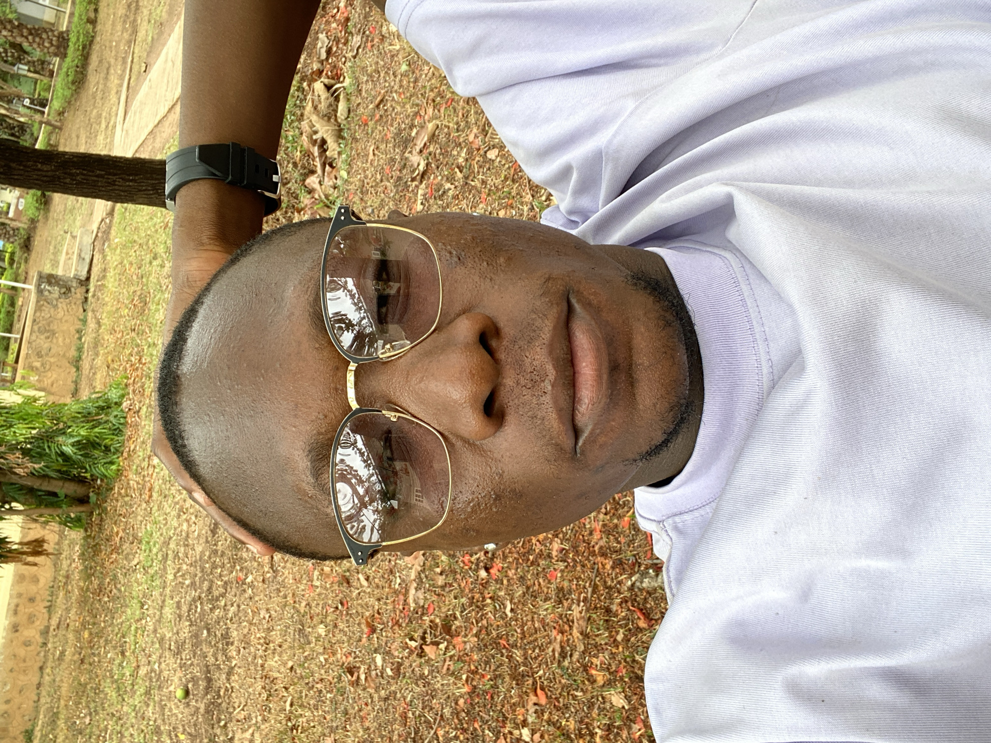 A selfie taken outdoors in Miango, Nigeria, shows a dark-skinned man wearing glasses and a white t-shirt. He has a black smartwatch on his left wrist. The background is dry grass and leaves, with trees and a building partially visible. The reflection in his glasses shows trees and sky.
