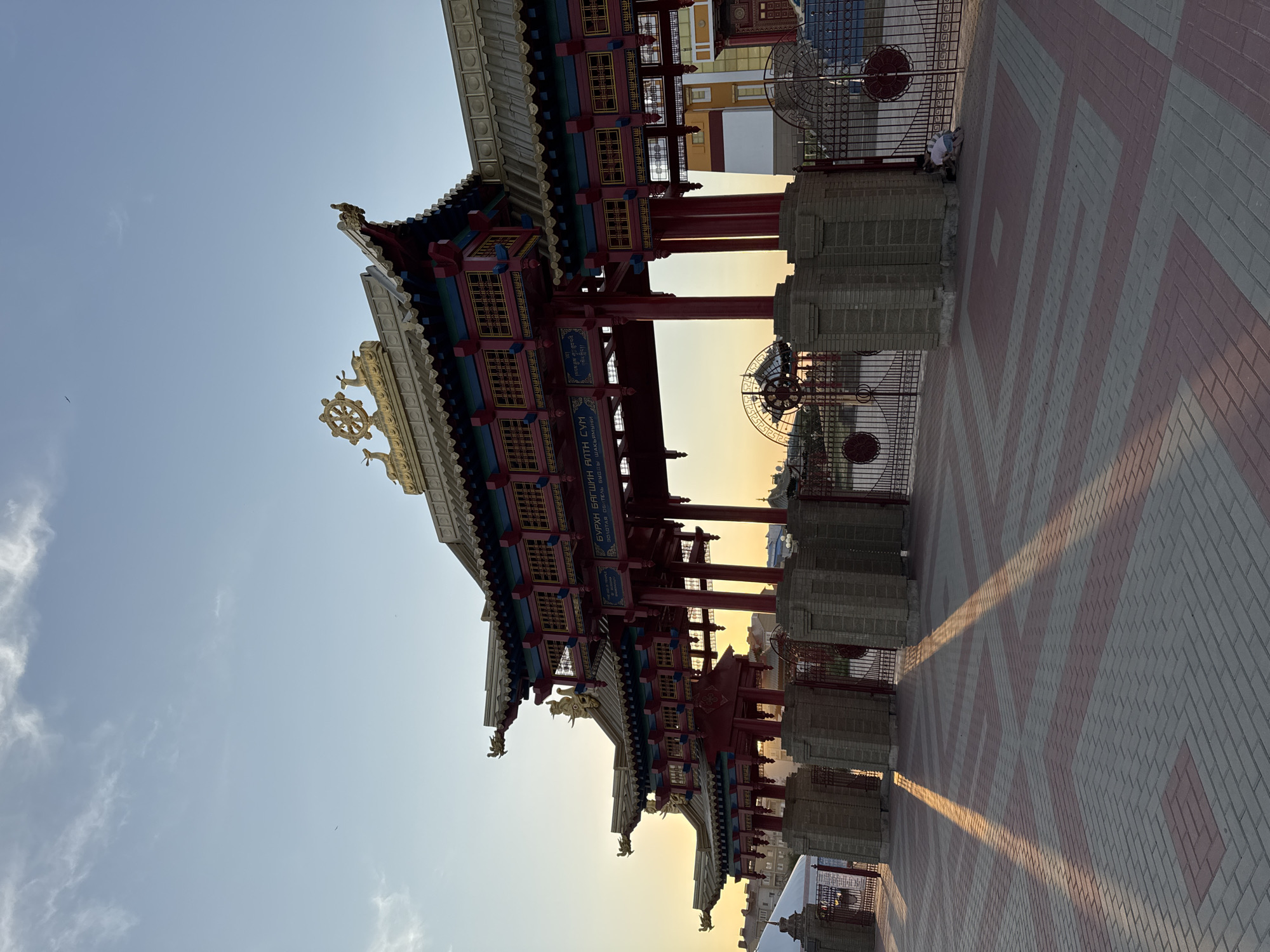 The image shows the Golden Abode of Buddha Shakyamuni, a Buddhist temple in Elista, Russia. The temple is a multi-tiered structure with red and gold accents, and traditional East Asian architectural elements such as curved roofs and ornate carvings. In the foreground, a patterned walkway made of red and grey bricks leads towards the temple. Sunlight casts long shadows across the walkway. Behind the temple, a clear blue sky with a few wispy clouds suggests it is late afternoon or early evening. The overall scene conveys a sense of grandeur and tranquility.