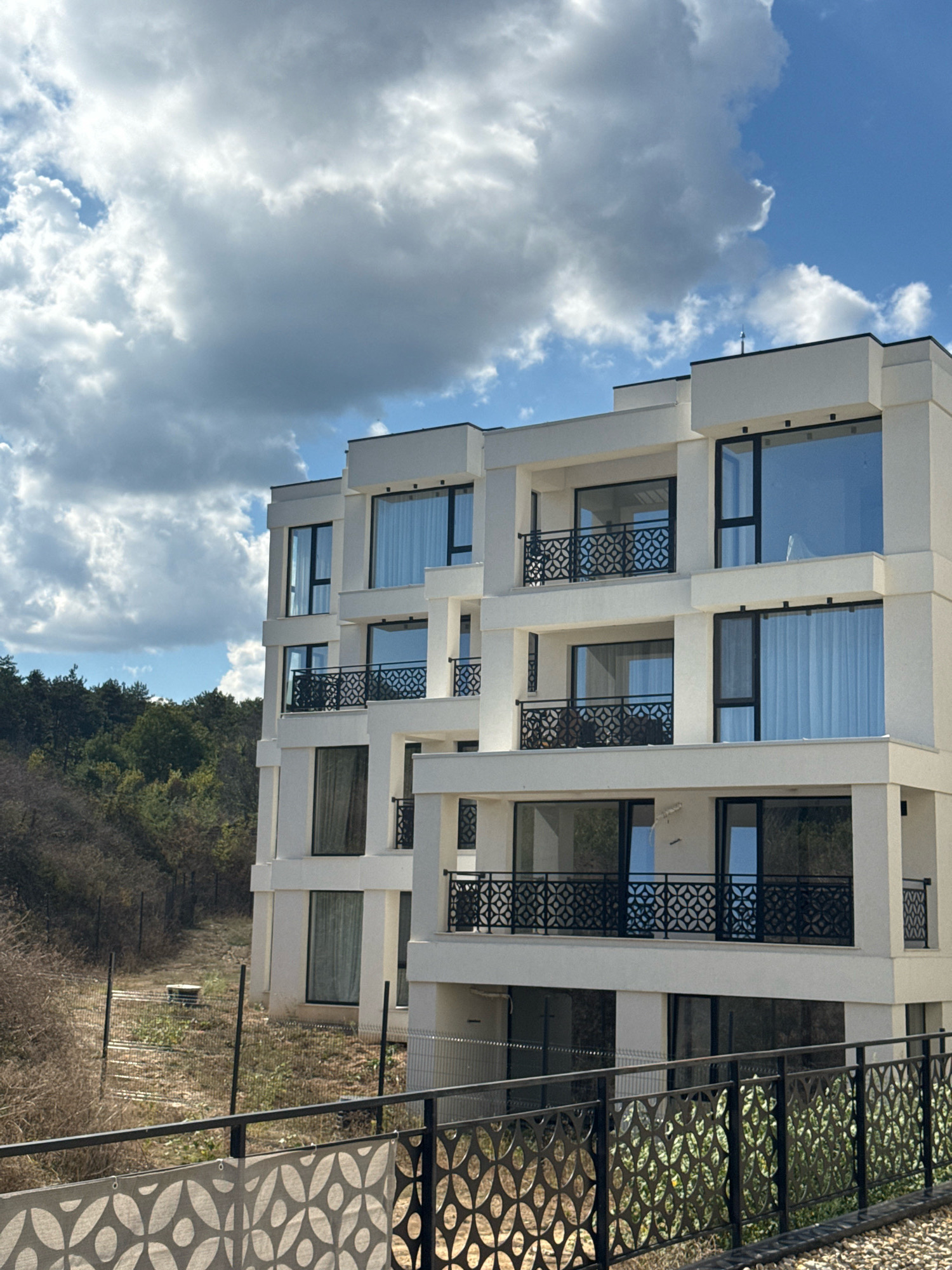 The image shows a modern, multi-story white building with dark-framed windows and balconies with decorative black railings. The building is situated in a natural, slightly overgrown environment with trees visible in the background. The sky is partly cloudy with white and gray clouds against a blue sky. The foreground features a metal fence with a decorative pattern. The scene appears to be a residential complex under construction or recently completed, located in the coastal city of Sozopol, Bulgaria, given the architectural style and the presence of lush vegetation. No people are visible in the image.