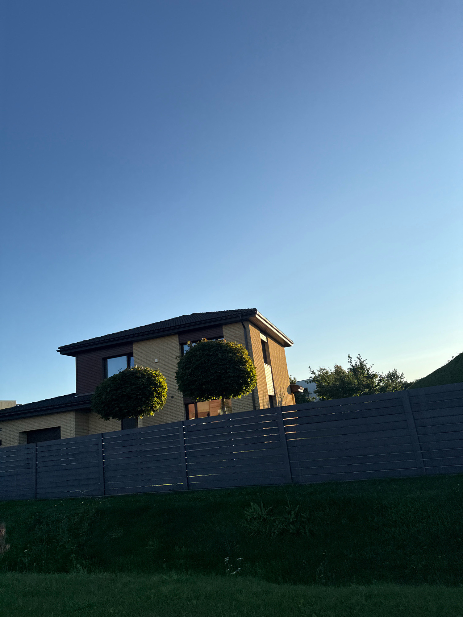 A modern, two-story house with beige brick and dark brown siding stands under a clear blue sky. The house features dark-framed windows and a dark roof. Two spherical, manicured trees are positioned in front of the house. A dark gray horizontal slatted fence runs across the foreground, with lush green grass visible behind it and below. The setting appears to be in a suburban area, likely Peetri alevik, Estonia, given the architectural style and landscaping.