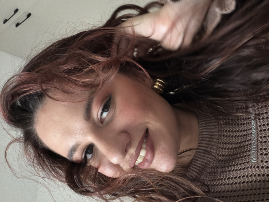 The image captures a close-up portrait of a young woman with long, wavy dark brown hair, accentuated with reddish-auburn highlights. Her head is tilted, and she is smiling broadly, revealing her teeth, while looking directly at the viewer with warm brown eyes. She wears noticeable eye makeup, including winged eyeliner, and has rosy blush on her cheeks and a natural lip color. A prominent feature is her gold-colored, multi-layered hoop earring on her visible ear. Part of her hand, adorned with a ring, is visible near her head, gently touching her hair.
She is dressed in a light brown, knitted sweater with a textured, open-knit pattern. The background is a plain, light-colored surface, likely an interior wall or ceiling, with two dark, curved fixtures visible in the upper left corner. The lighting appears bright and even, indicative of an indoor setting during the daytime. The woman's expression conveys happiness, friendliness, and contentment, suggesting a casual and cheerful moment, possibly a selfie. The visible text "MONOCHROM" is partially displayed vertically along the right side of her sweater. The scene is contextualized as being in Budva, Montenegro, although no specific outdoor location cues are visible within the frame.