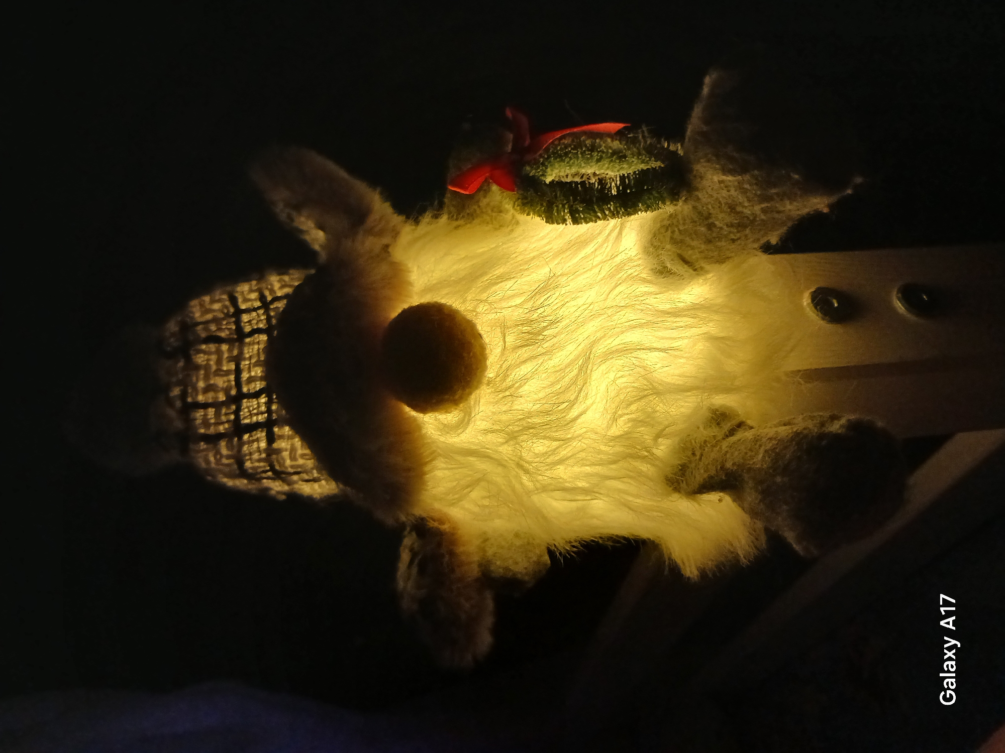 The image shows a stuffed gnome-like figure with a glowing, fluffy body and a textured hat. It has a round nose and a small wreath with a red ribbon on its head. The figure appears to be sitting on a light-colored wooden surface with visible screws. The background is dark. There is no indication that this image was taken in Yekaterinburg, Russia.