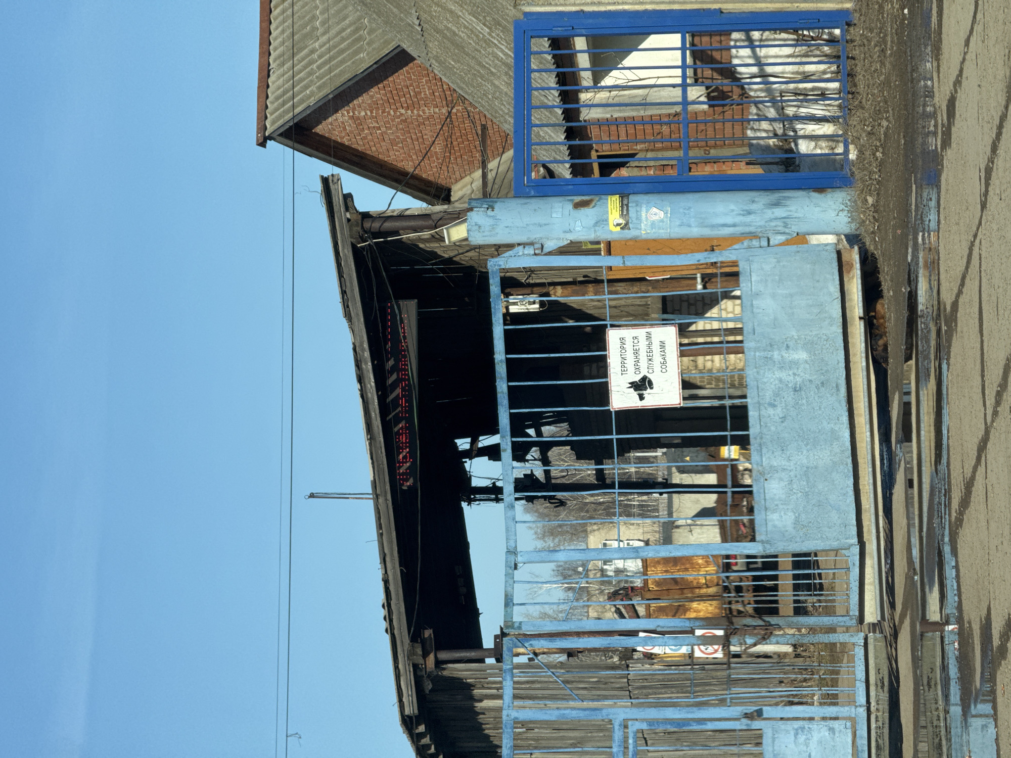 An outdoor scene captures the entrance to what appears to be an industrial or storage area, likely located in the city of Chaikovsky, Russia. The entrance is marked by a dilapidated, blue metal gate, with the top section having a square grid pattern. A sign in Russian with a dog icon is affixed to the gate, translating to "The territory is guarded by service dogs."
Behind the gate, one can see a partially open building with a dark interior, suggesting neglect or abandonment. The roof of the building is a mix of old roofing tiles and what seems to be exposed brick. A portion of a neighboring building can also be seen.
To the right of the gate, there's a paved road showing signs of wear, with tire tracks and some sort of liquid stain visible on the surface. The overall impression is that of a neglected or industrial area in a Russian urban setting. The sky above is clear and blue.