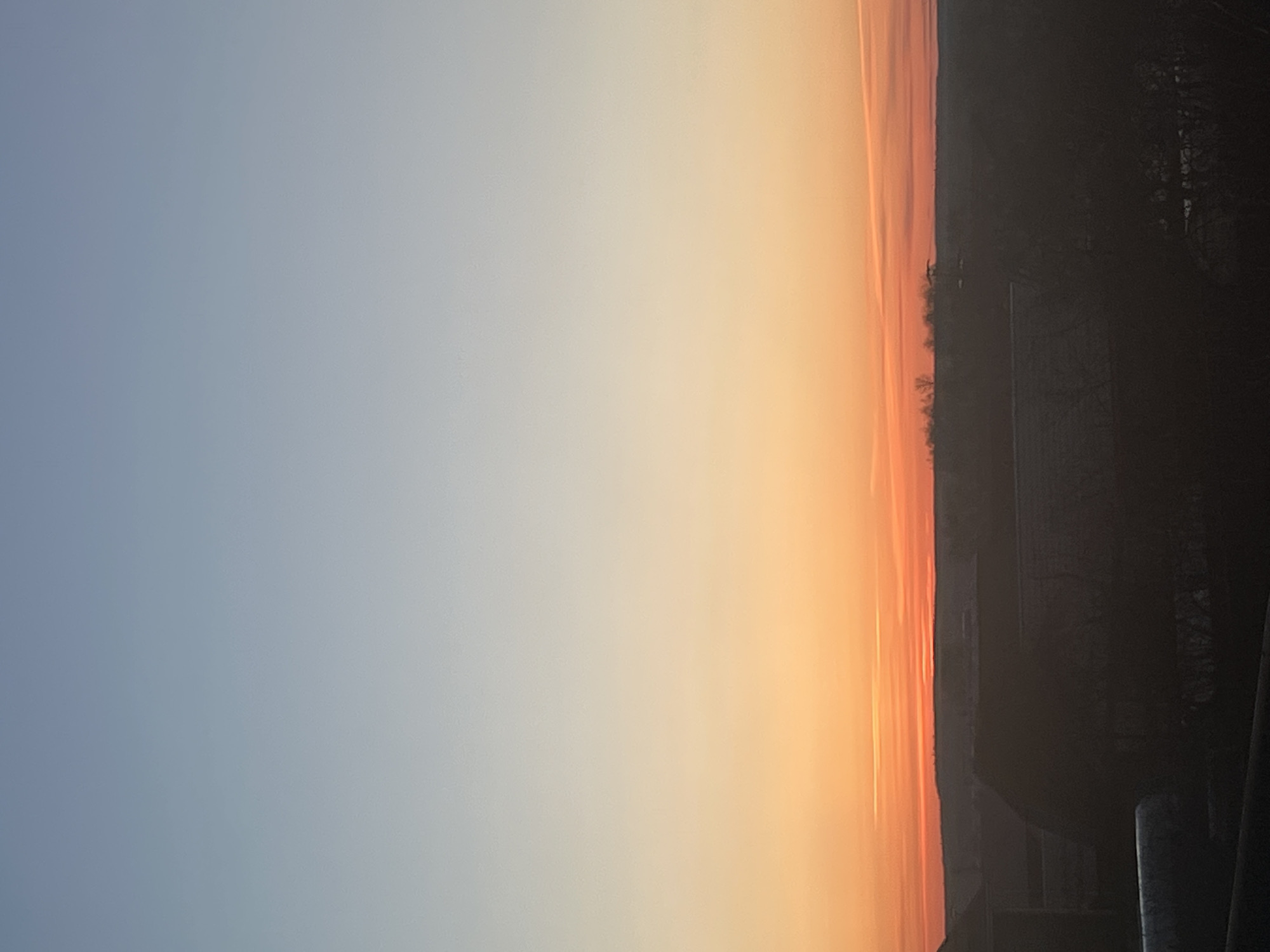 The image shows a view of the sky at sunset. The sky transitions from a pale blue on the left to orange and yellow on the right. A dark silhouette of trees and buildings is visible on the right edge. The location in the city of Votkinsk, Russia, cannot be determined from the image.