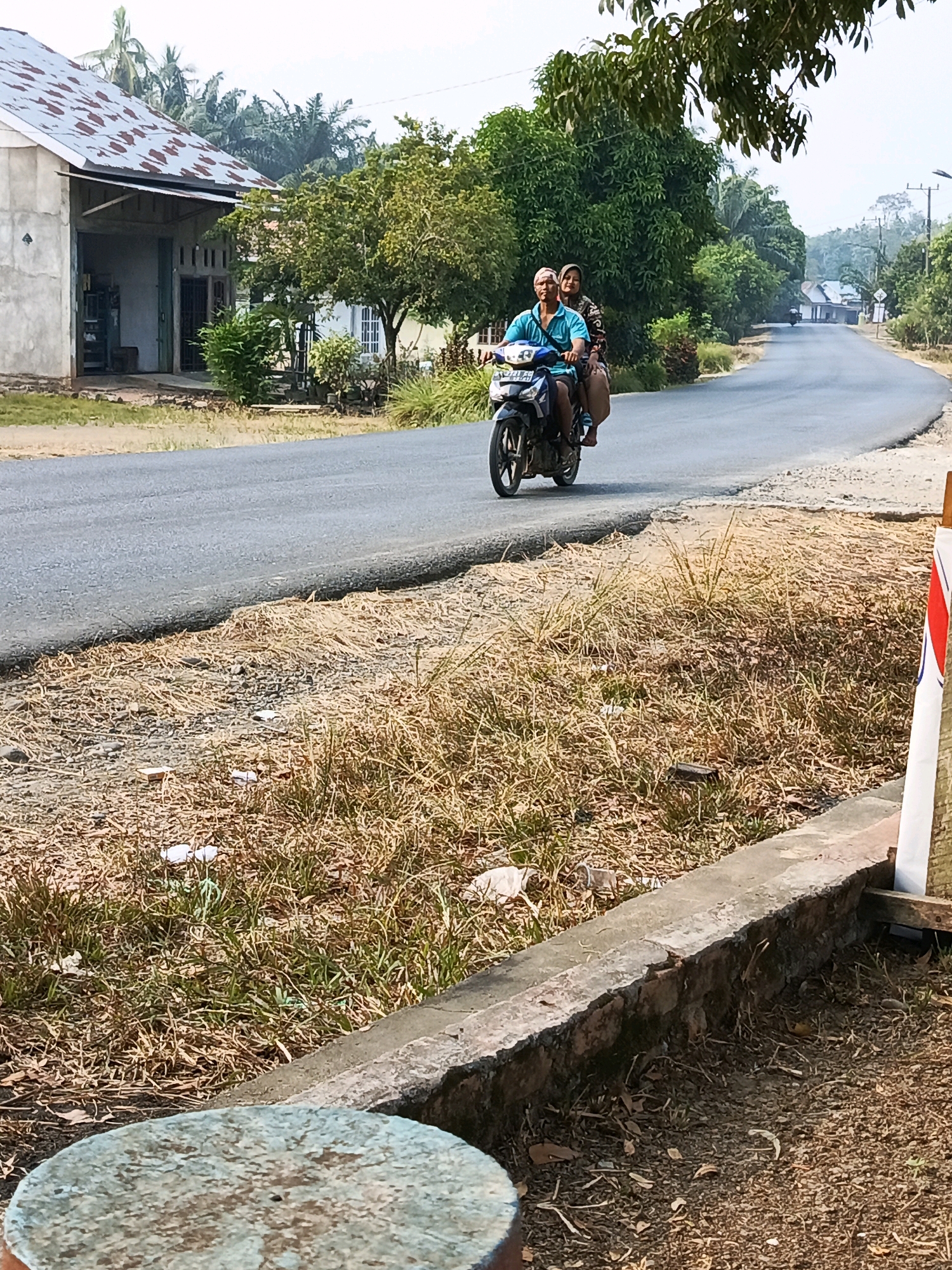 A man and a woman ride a motorcycle on a paved road in a rural area. The man is driving and the woman is a passenger. Both are wearing casual clothing. The motorcycle is black and blue. The road is flanked by dry grass and trees. In the background, there are houses and power lines. The sky is hazy. The scene appears to be in Wonosari, Indonesia.