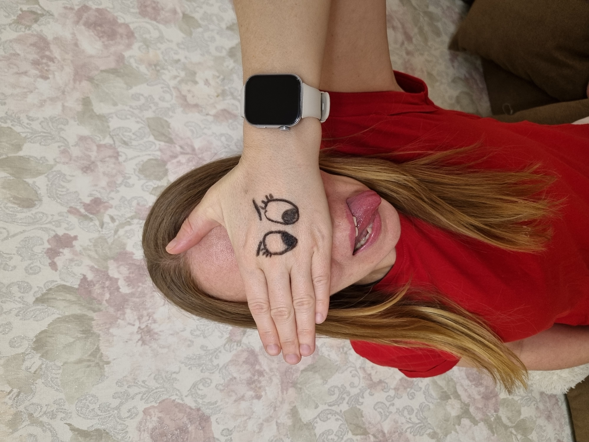 The image captures a person lying down on a patterned surface, likely a bed, in an indoor setting in Chaikovsky, Russia. The subject, appearing to be a woman with long, light brown hair, is wearing a red t-shirt. Her right hand is positioned over her face, covering her eyes. On the back of this hand, two cartoon-style eyes are drawn with a black marker, creating a humorous effect. A light-colored smartwatch with a dark, unlit screen is visible on her right wrist. Beneath her hand, her mouth is slightly open, and her tongue is playfully sticking out, contributing to a silly and lighthearted expression. The surface she is lying on is a light-colored fabric featuring an intricate floral pattern in muted tones of pink, grey, and white. In the upper right corner of the frame, a dark brown pillow or cushion is partially visible. The scene suggests a relaxed and playful moment, with the time of day being indeterminate. No other explicit text is visible besides the drawn eyes.