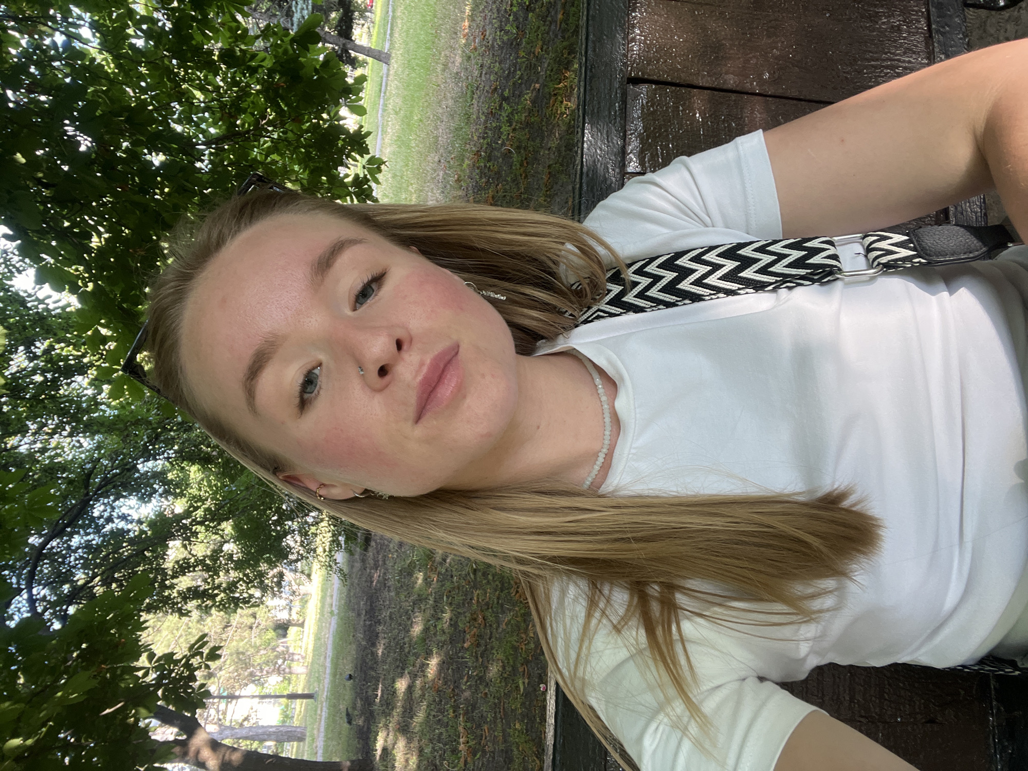 This is a selfie of a young woman with blonde hair and blue eyes, taken outdoors. She is wearing a white t-shirt and a necklace. A black and white patterned strap is visible across her chest, likely from a bag. Her face is turned towards the camera, and she has a neutral expression. The background shows greenery, including trees with dappled sunlight filtering through the leaves, and a grassy area that appears to be a park. There is also a dark wooden surface, possibly a bench, on which she is seated or leaning. The lighting suggests it is daytime, likely sunny. The overall atmosphere is casual and relaxed, hinting at an outdoor leisure activity. There is no visible text in the image.