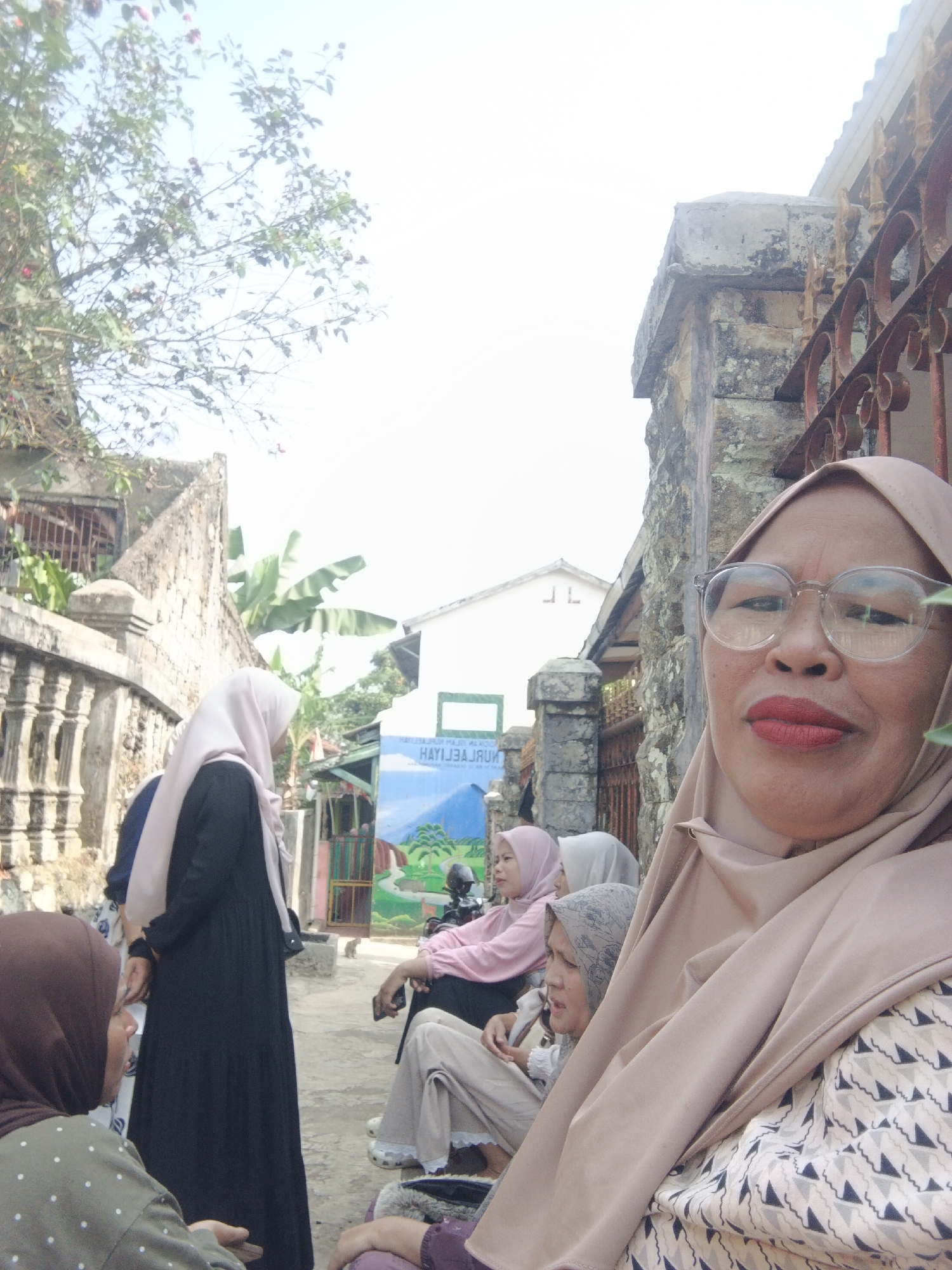 In Warungkiara, Indonesia, a group of women are gathered outdoors. The woman in the foreground, facing the camera, wears a tan hijab and glasses, and has red lipstick. She is close to the camera. Behind her, several other women are seated and standing, also wearing hijabs. One woman in a black dress and tan hijab stands facing away from the camera. Another woman in a pink hijab and white headscarf is seated. A woman in a grey headscarf is also seated. In the background, there is a mural depicting a landscape with mountains and a blue sky. The environment appears to be a street or courtyard, with a stone wall on the right and a lower wall with decorative pillars on the left. Trees and foliage are visible, and a white building is in the distance. The lighting suggests it is daytime.