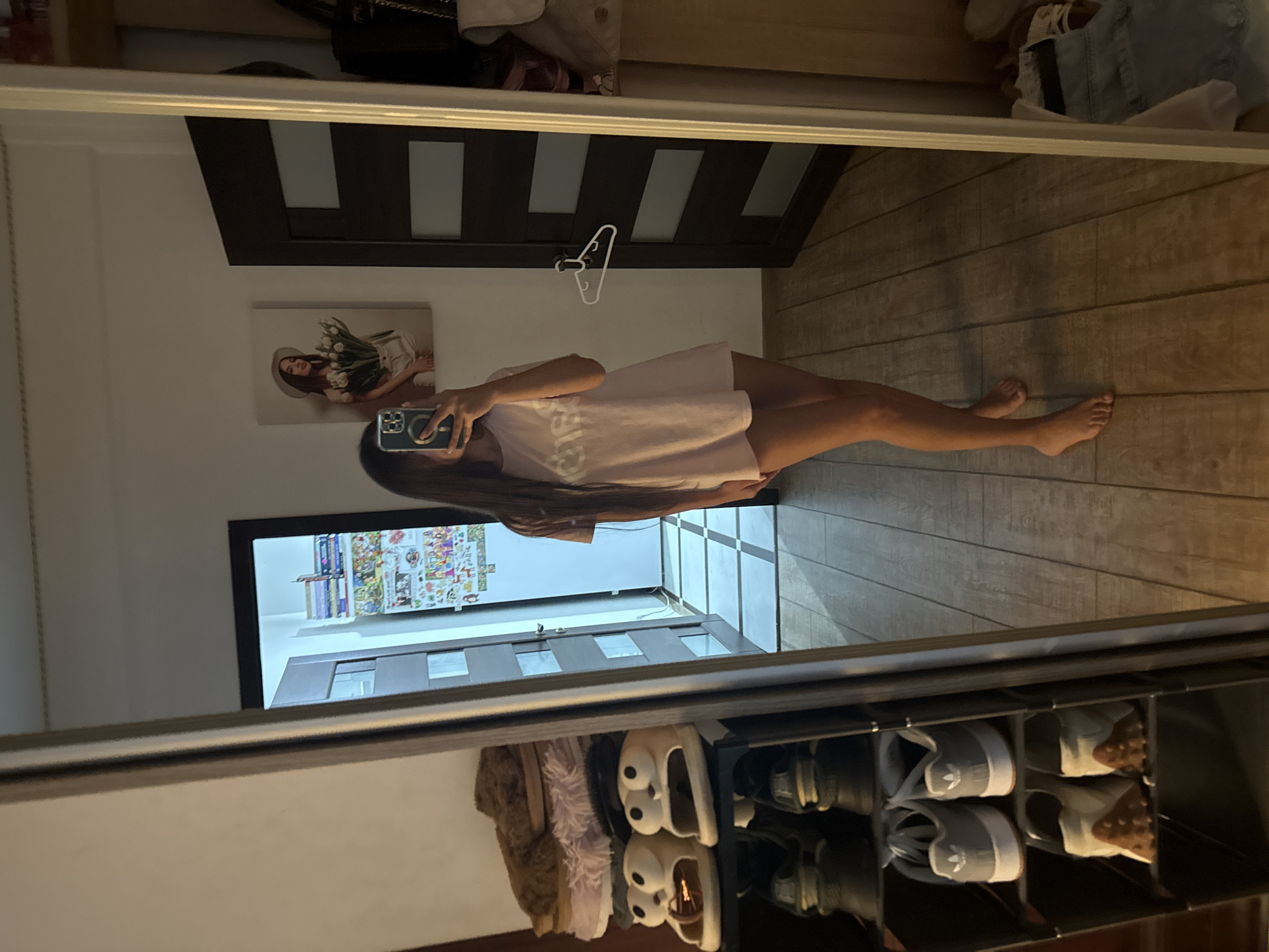 In an interior residential setting, a young woman stands in front of a full-length mirror, taking a selfie with her smartphone. She is wearing a light pink, oversized t-shirt and appears to be barefoot. The mirror reflects a room with wooden flooring and white tiled sections. To the left, a multi-tiered shoe rack is visible, holding various footwear, including fuzzy slippers, sandals shaped like cartoon characters, and athletic shoes. Further to the left, a doorway leads into another area of the home. On the wall in the background, there is a framed artwork depicting a woman in a hat holding a bouquet of tulips, and a dark-colored door with horizontal glass panels. A white hanger is attached to this door. The scene suggests a casual moment in a private residence, potentially in Chișinău, Moldova, given the style of the interior and the presence of certain decorative elements common in the region.