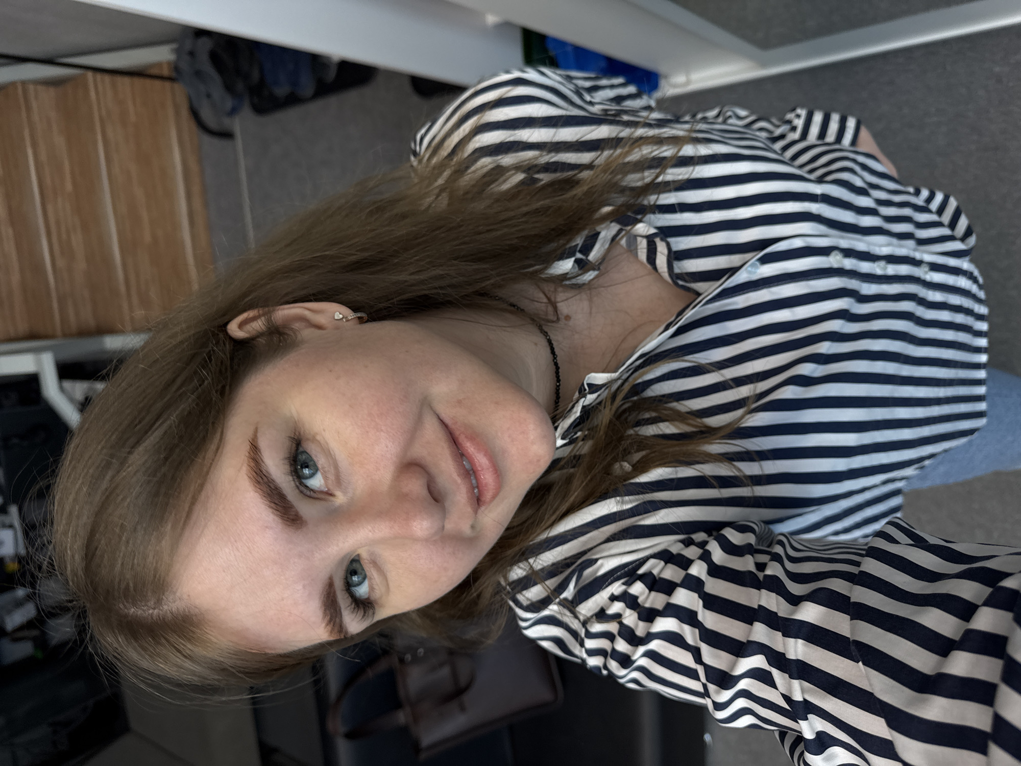 A woman with light brown hair and blue eyes is posing for a photo. She is wearing a striped shirt with black and white lines and a black necklace. She has a small earring in her left ear. The background is a mix of interior elements, including a wooden floor, a grey carpeted area, a white door, and some furniture and personal belongings partially visible. The location appears to be indoors, possibly in an apartment or home setting in Perm, Russia. The photograph is taken at an angle, with the woman looking up at the camera.