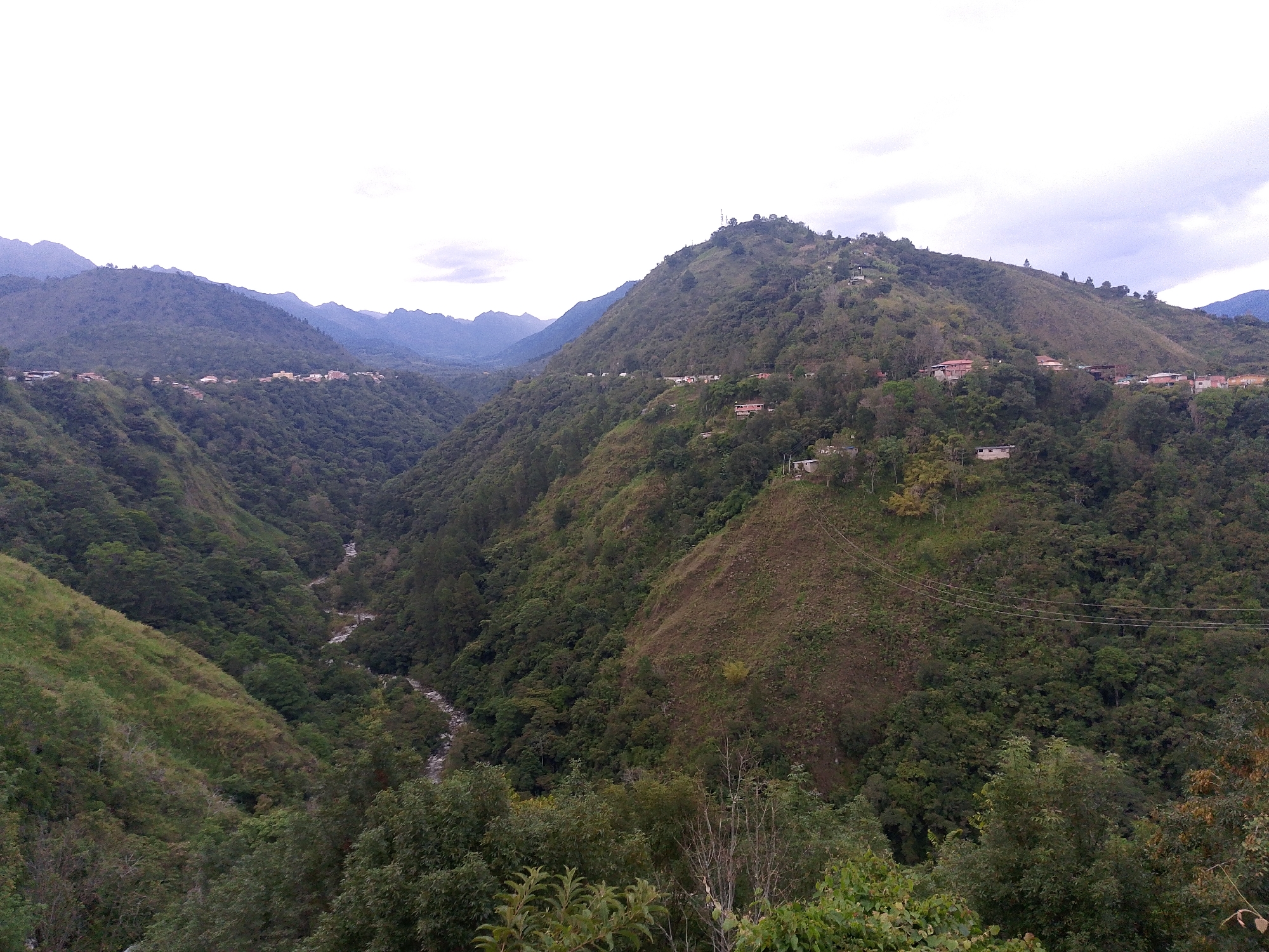 The image shows a mountainous landscape in the city of Mérida, Venezuela. Lush green vegetation covers the slopes, with a river flowing through a deep valley. Clusters of houses are scattered along the hillsides, suggesting a rural or semi-rural settlement. The sky is overcast, casting a soft light on the scene. Power lines are visible stretching across the mountainside.