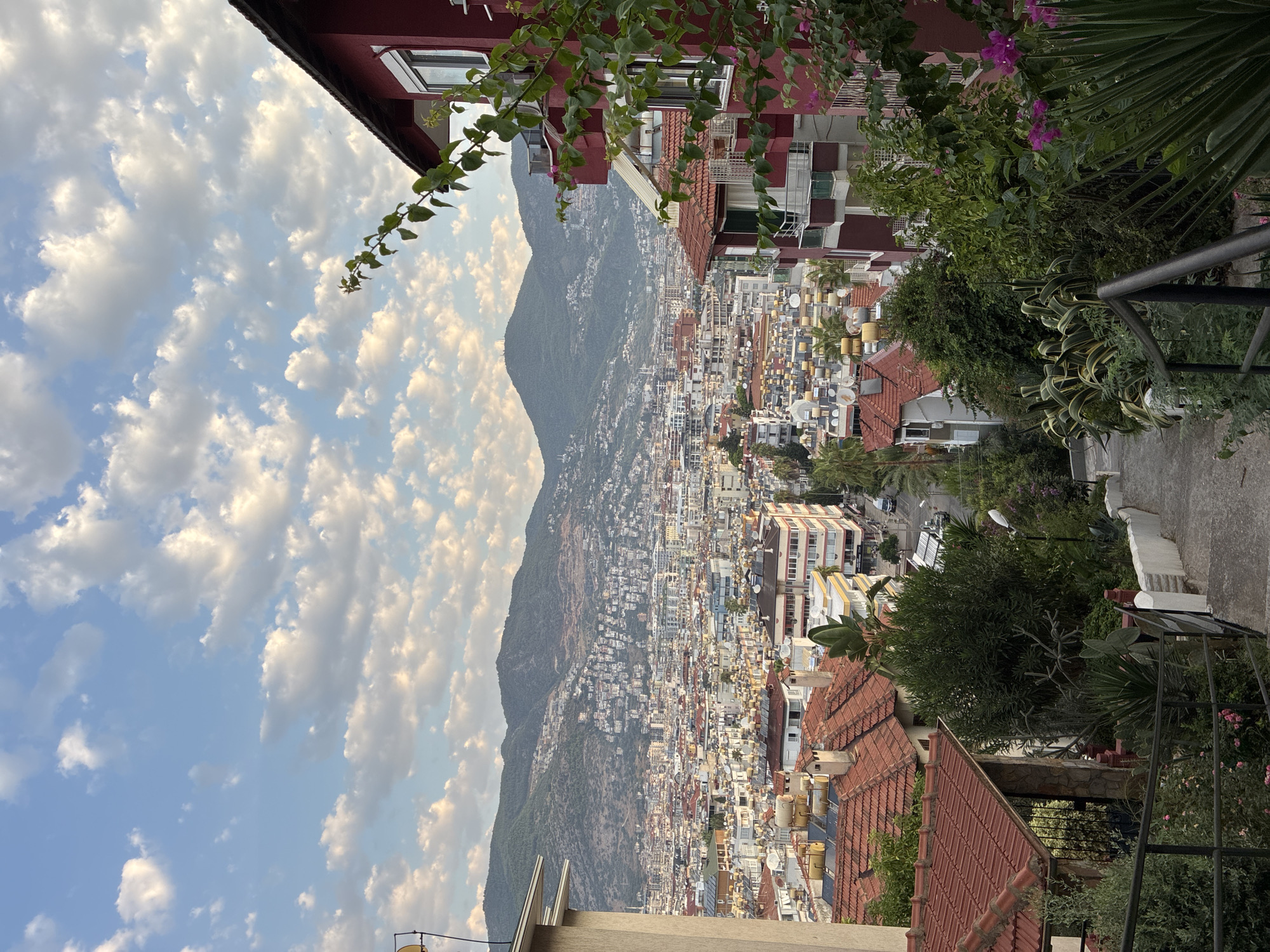The image captures a panoramic view of Alanya, Turkey, from an elevated vantage point. The foreground features a descending concrete staircase bordered by lush green foliage and flowering plants. A dark metal railing runs along the side of the stairs. To the right, a multi-story building with red and white facades is partially visible, with a vine trailing down its side.
In the midground, the dense urban landscape of Alanya unfolds. Numerous buildings with red-tiled roofs dominate the scene, interspersed with apartment blocks of varying architectural styles and colors, including white, beige, and red. Many rooftops are equipped with solar water heaters, a common sight in the region. A street with a few parked cars is visible winding through the city.
The background is characterized by rolling, green mountains that form a dramatic backdrop to the city. The sky above is a clear blue, dotted with scattered, fluffy white clouds, suggesting pleasant weather. The overall impression is one of a vibrant coastal city nestled against a mountainous terrain.