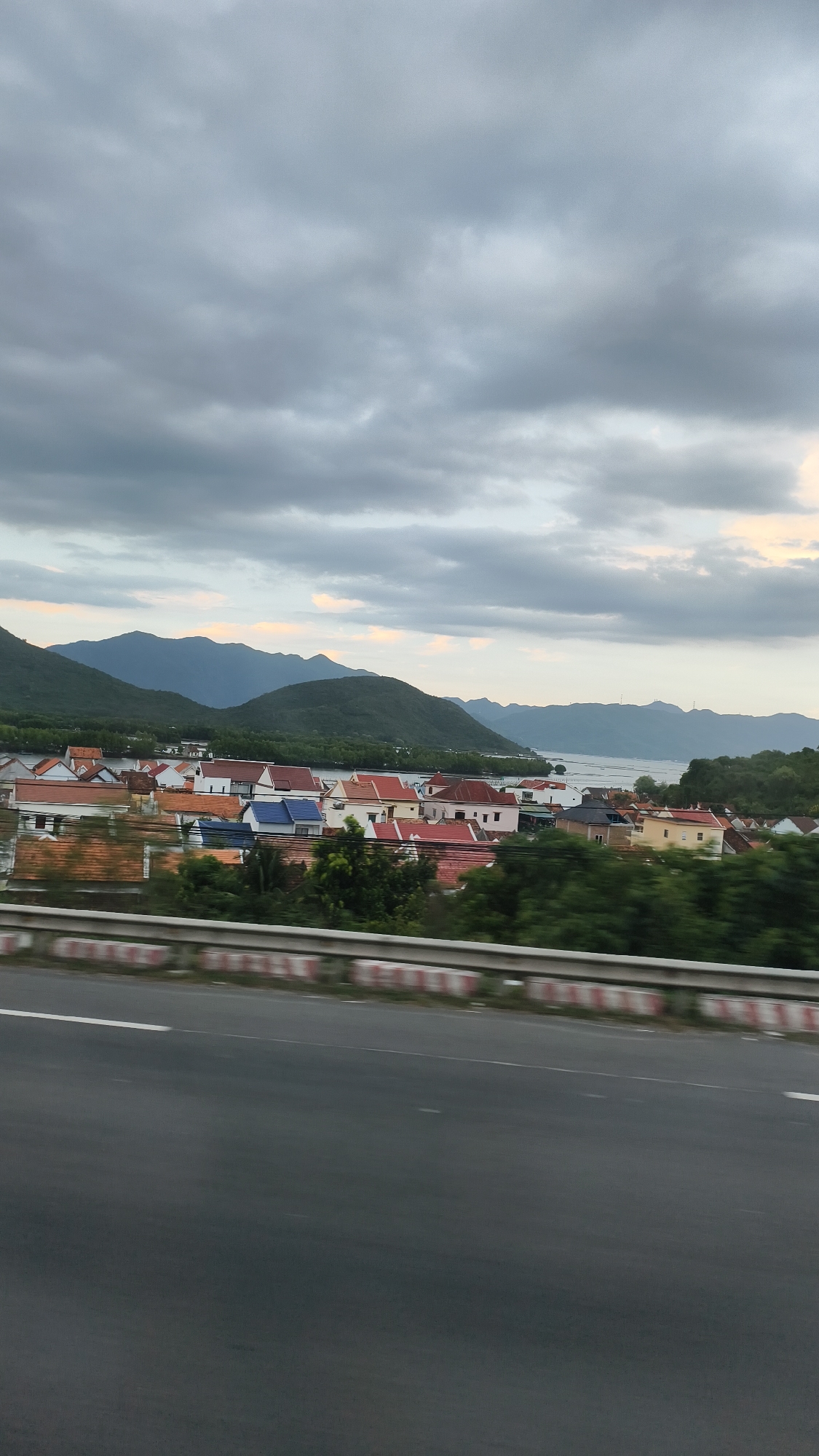 A telephoto shot captures a residential area situated between a body of water and a mountain range under a cloudy sky. The foreground features a road with a concrete barrier along the side. Beyond the barrier, a cluster of houses with red, white, and blue roofs is visible, nestled amongst lush green trees. In the middle ground, a calm body of water stretches across, reflecting the muted tones of the overcast sky. The background is dominated by rolling green mountains, their peaks softened by distance and the atmospheric haze. The overall scene depicts a tranquil coastal or lakeside community.