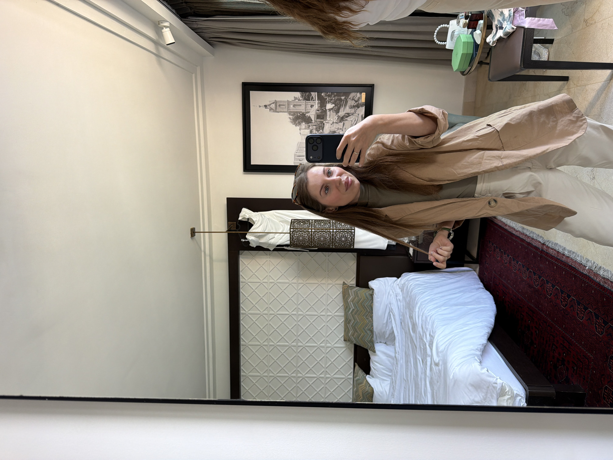 In a mirror selfie, a young woman with long brown hair, wearing a light brown jacket over a green top and light-colored pants, holds a dark-colored smartphone to capture her reflection. Sunglasses rest on top of her head, and a ring is visible on her left ring finger. She has a relaxed expression with a subtle smile.
The setting appears to be a bedroom, possibly in a hotel or apartment in Tel-Aviv, Israel. Reflected in the mirror behind her is a neatly made bed with crisp white bedding and a dark wooden headboard featuring a geometric patterned panel. A red, ornate, traditional-style rug lies on the floor beside the bed. On the wall above the bed, a large framed black and white artwork depicts an old building or townscape, possibly a historical landmark. To the left, a track light fixture is visible on the ceiling, illuminating the room. A small, dark, ornate box sits on a bedside table. In the upper right corner of the reflection, a small table or shelf holds a white handbag, a green octagonal box, a smaller mirror, and various personal items. The walls are light-colored with simple molding, contributing to a clean and sophisticated aesthetic.