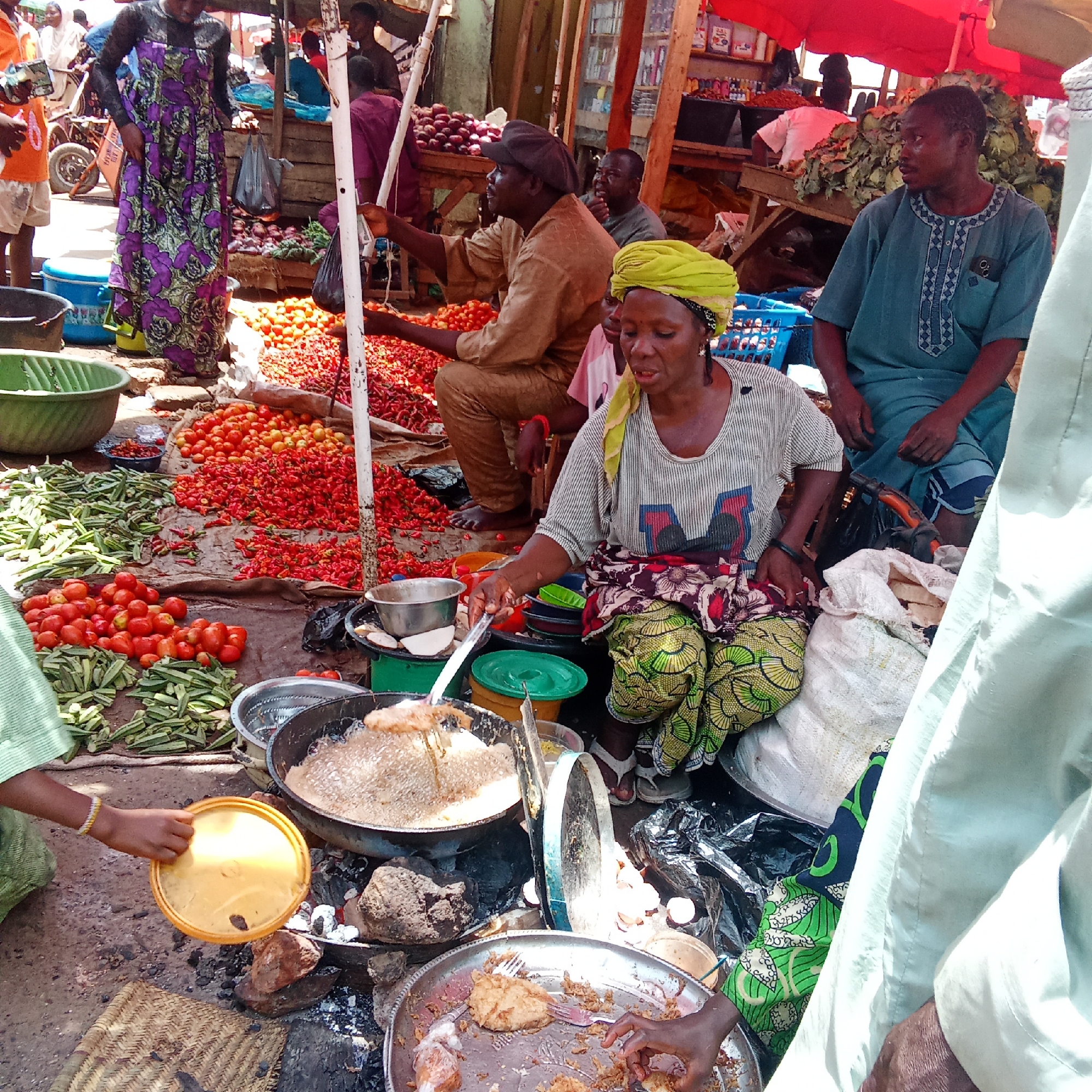 The image depicts a vibrant street scene in Akwanga, Nigeria, likely a marketplace. At the forefront, a woman wearing a patterned shirt, head wrap, and colorful pants is actively cooking; she is using a spatula to tend to food frying in a large pan over an open fire built on the ground. Beside the fire, a tray with what appears to be cooked or fried food is present, with hands reaching in.
Behind the cooking area are displays of fresh produce: piles of tomatoes, okra, and red peppers. Men are sitting among the produce, one wearing a brown robe and another a blue embroidered shirt. Overhead, there's a partial view of market stalls with red awnings. People are milling about in the background. The general scene is bustling with activity and trade.