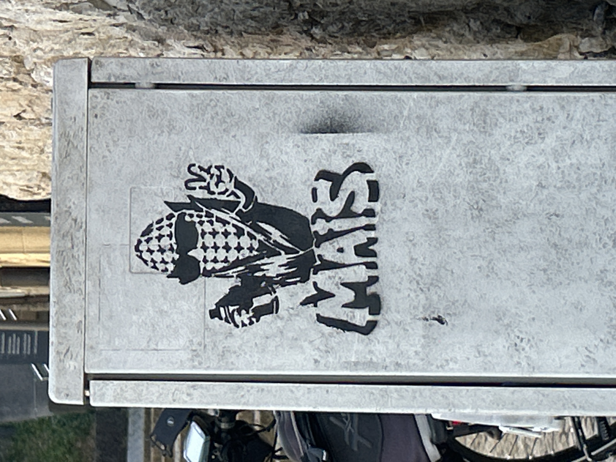 An outdoor, eye-level, medium shot shows graffiti on a utility box. The graffiti is in black on a textured gray background. At the top of the box, a stencil depicts a cob of corn wearing sunglasses and a checkered bandana. The cob is holding a spray can in its right hand and making a peace sign with its left. Below the cob, the word "MAIS" is stenciled in large, blocky letters. A bicycle with a black bag on the back is partially visible to the left of the utility box, and a stone wall is to the right. The scene is located in Bad Friedrichshall, Germany.