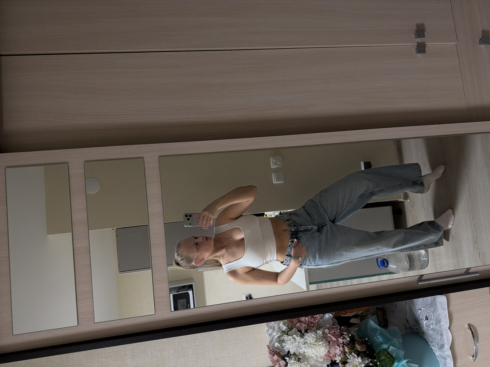 A young, blonde woman is taking a selfie in front of a wardrobe with mirrored doors. She is wearing a white crop top and light-wash jeans. The reflection shows a room with light-colored walls, a microwave, a bouquet of flowers in a blue vase, and a water bottle on the floor. The mirrored doors appear to be part of a wardrobe system. No evidence indicates it is in Izhevsk, Russia.