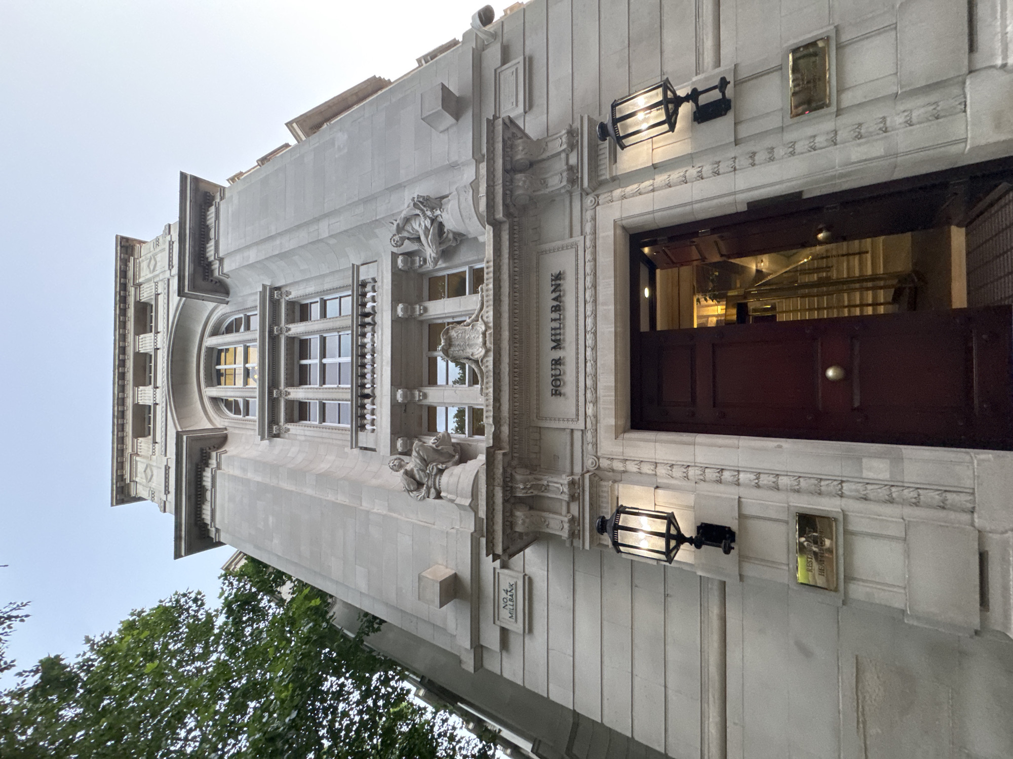 The image shows the entrance to the building at Four Millbank in London, United Kingdom. The building is constructed of light-colored stone and features ornate architectural details, including sculptures above the entrance and decorative moldings. Two large black lanterns with illuminated bulbs are mounted on either side of the main doorway. The entrance itself is an imposing set of dark red double doors, one of which is ajar, revealing a brightly lit staircase inside. Above the doors, the name "FOUR MILLBANK" is carved into the stone facade. To the left of the entrance, a sign reads "NO. 4 MILLBANK". To the right of the entrance, a brass plaque with text that appears to read "RESTAURANT HEATHERCOURT" is visible. A large tree with green foliage is present on the left side of the frame, partially obscuring the building. The sky above is a pale blue, suggesting it is either early morning or late evening.