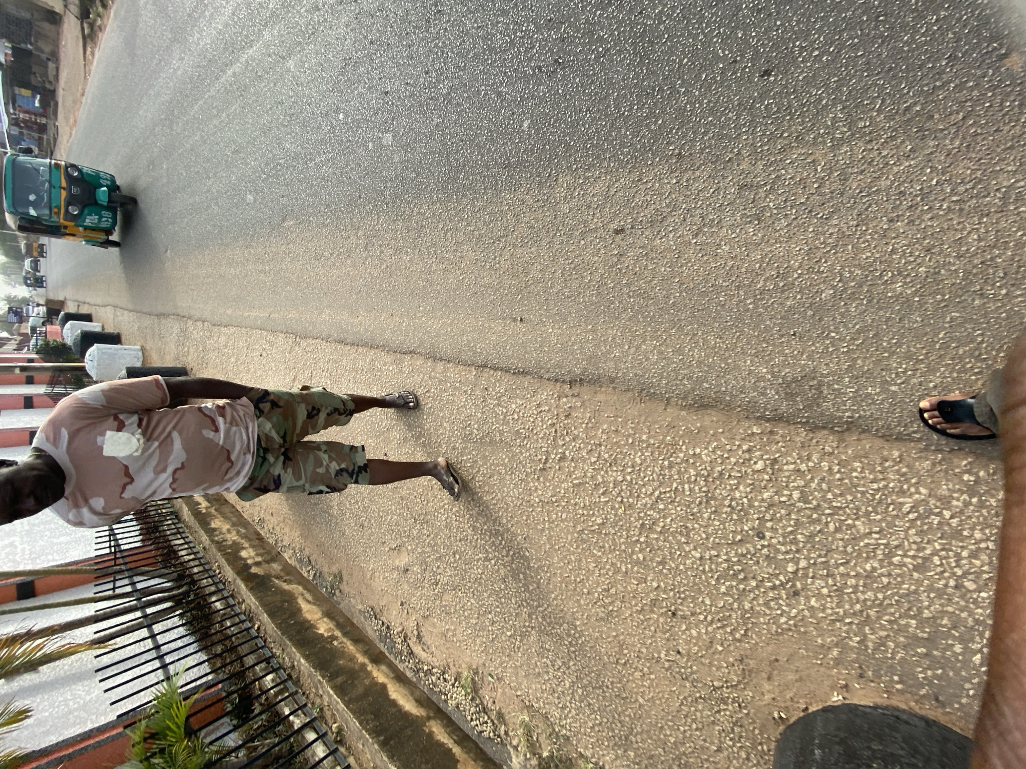 An outdoor, eye-level shot captures a street scene in Bukuru, Nigeria, under bright daylight. The image is oriented vertically, showing a man standing on the dirt shoulder beside a paved road. He wears a camouflage-patterned t-shirt and shorts, with flip-flops on his feet.
Further down the road, a green and yellow auto-rickshaw, commonly known as a "Keke Napep" in Nigeria, is visible. Text on the back of the auto-rickshaw includes "49R" and "1938". Other distant vehicles can be seen on the road.
To the left of the standing man, a black metal fence runs along the roadside, partially obscured by some green foliage. White and black bollard-like structures are positioned behind the fence. The road surface appears to be asphalt, with a lighter-colored, coarse-textured shoulder. The foot of another person, likely the photographer, wearing black sandals, is visible in the bottom right corner of the frame. The clear lighting suggests a sunny day. No strong emotions are discernible from the subjects.