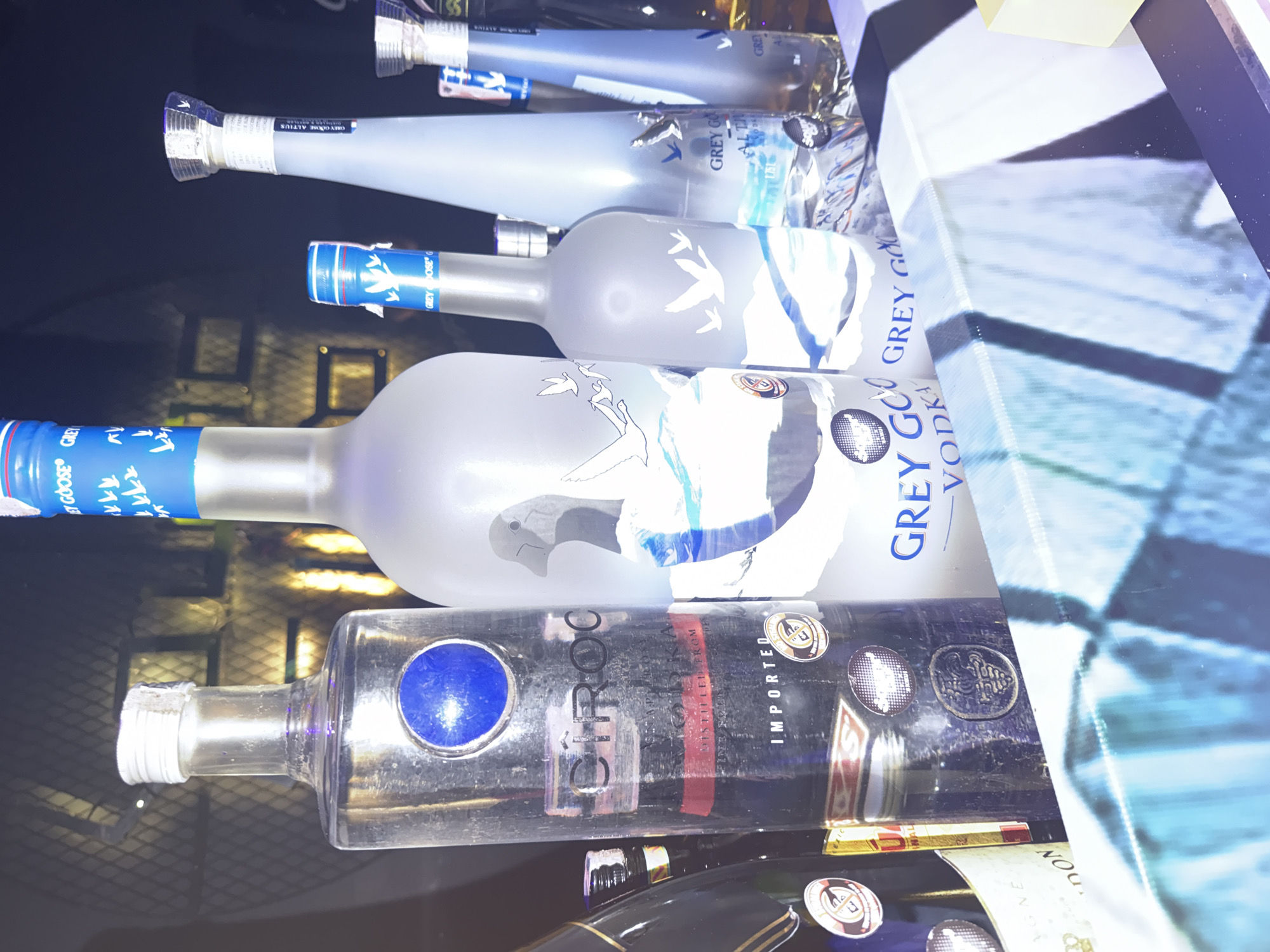 The image shows a selection of liquor bottles on a reflective surface, likely a bar top. There are multiple bottles of Grey Goose vodka, identifiable by the brand name and goose imagery on the labels. Also present is a bottle of Cîroc vodka, distinguished by its dark bottle and prominent logo. The bottles are arranged in a row, with some overlapping others, suggesting they are on display or in storage behind a bar.
The image does not contain sufficient details to pinpoint the specific location within บ้านบางรักษ์, Thailand.