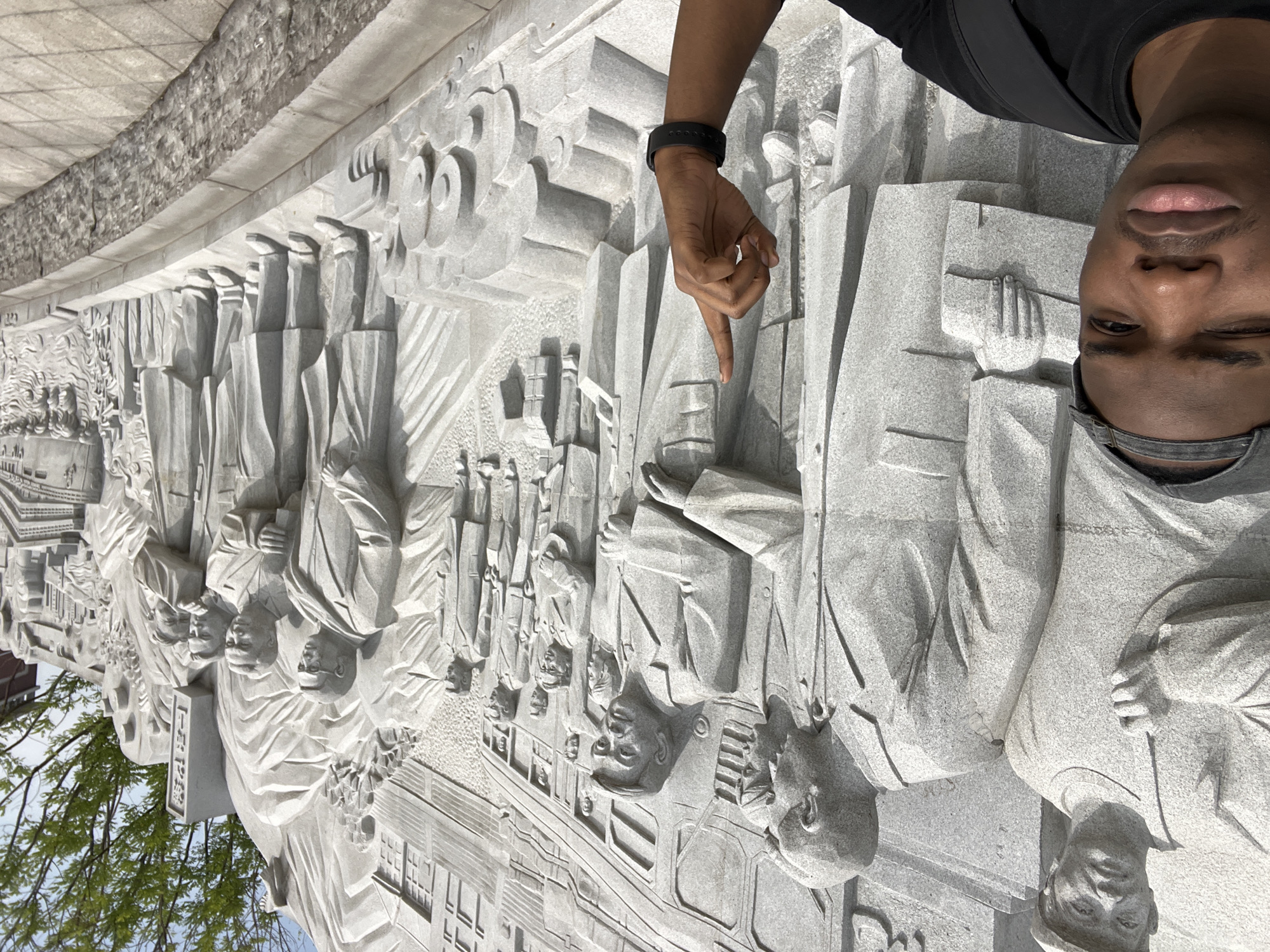 A person stands next to a large, gray stone carving in Chongqing High-tech Zone, China. The stone carving depicts a relief of people and architectural elements. The person points to the carving with their right hand, wearing a black fitness tracker on their wrist. The person is Black, with dark hair, brown eyes, and pink lips. They are wearing a black shirt and a gray baseball cap, and a black bag strap is visible across their shoulder. The stone carving is highly detailed, with depictions of figures in period clothing and a stylized depiction of buildings. The background features a sky with trees visible. The carving is set against a light gray, tiled surface.