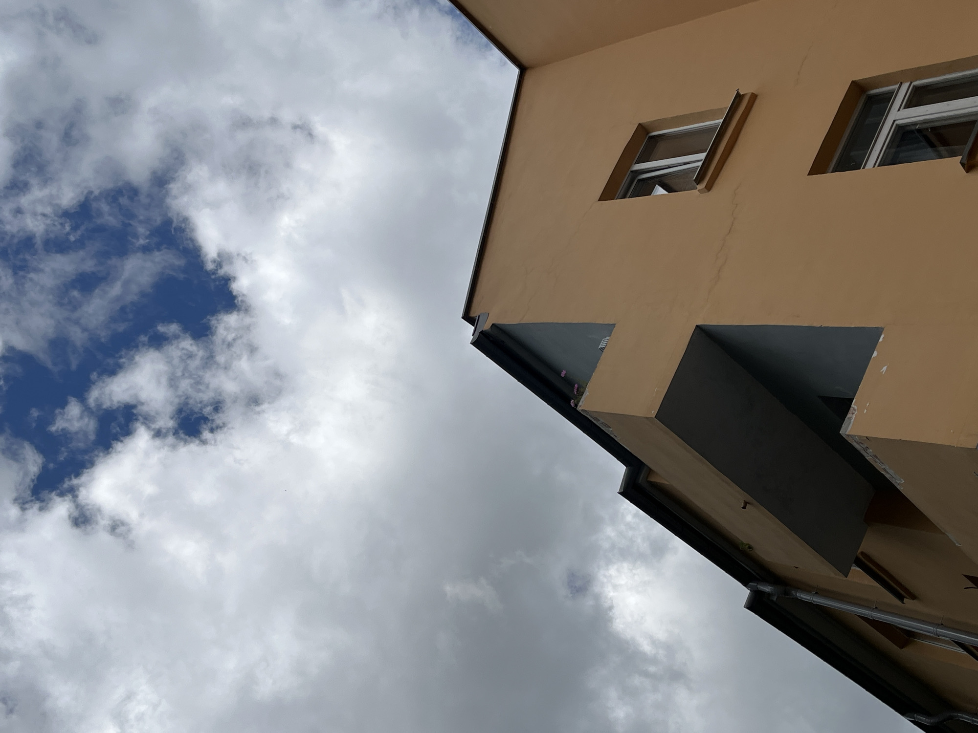 The image shows the upper floors of an apartment building with an orange facade and a dark roof. The building has several windows and balconies, some of which are recessed and have grey walls. The sky is partially cloudy, with patches of blue sky visible through white and grey clouds. The image is taken from a low angle, looking upwards at the building and the sky. There are no people visible in the image. The architecture suggests a European urban environment. No specific landmarks are visible that would allow for a definitive identification of the location within Vitsebsk, Belarus.