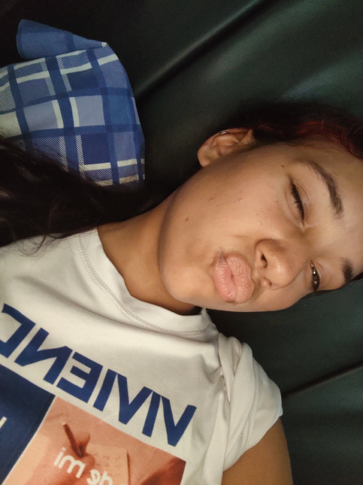 In Piloncito, Venezuela, a young woman is lying down with her eyes closed and puckering her lips. She has dark hair with hints of red, and is wearing a white t-shirt with blue text that appears to be "VIVENCIAS" printed on it. To her left, a blue and white plaid pillow is visible. The background is a dark, textured surface, likely a couch or chair.