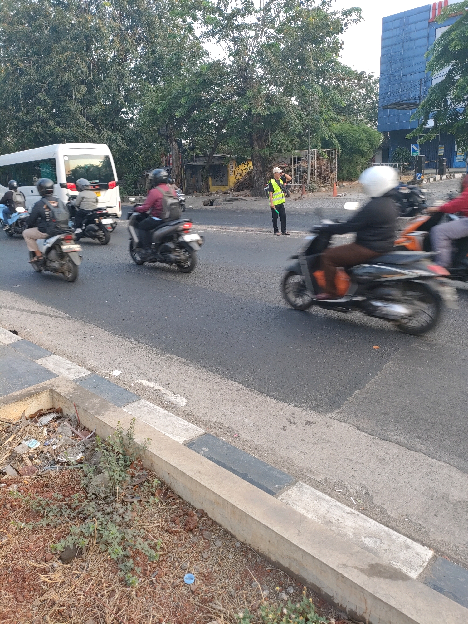 In Bekasi, Indonesia, on a street with a light gray asphalt surface, a person wearing a bright yellow safety vest and a white cap is standing on the side of the road. They are holding a black stick. Several motorcycles are on the road, with some riders in motion, evidenced by motion blur. One white van is visible in the background. The environment includes trees with green foliage and a blue building. There is a concrete curb along the side of the road with some dry vegetation and debris.