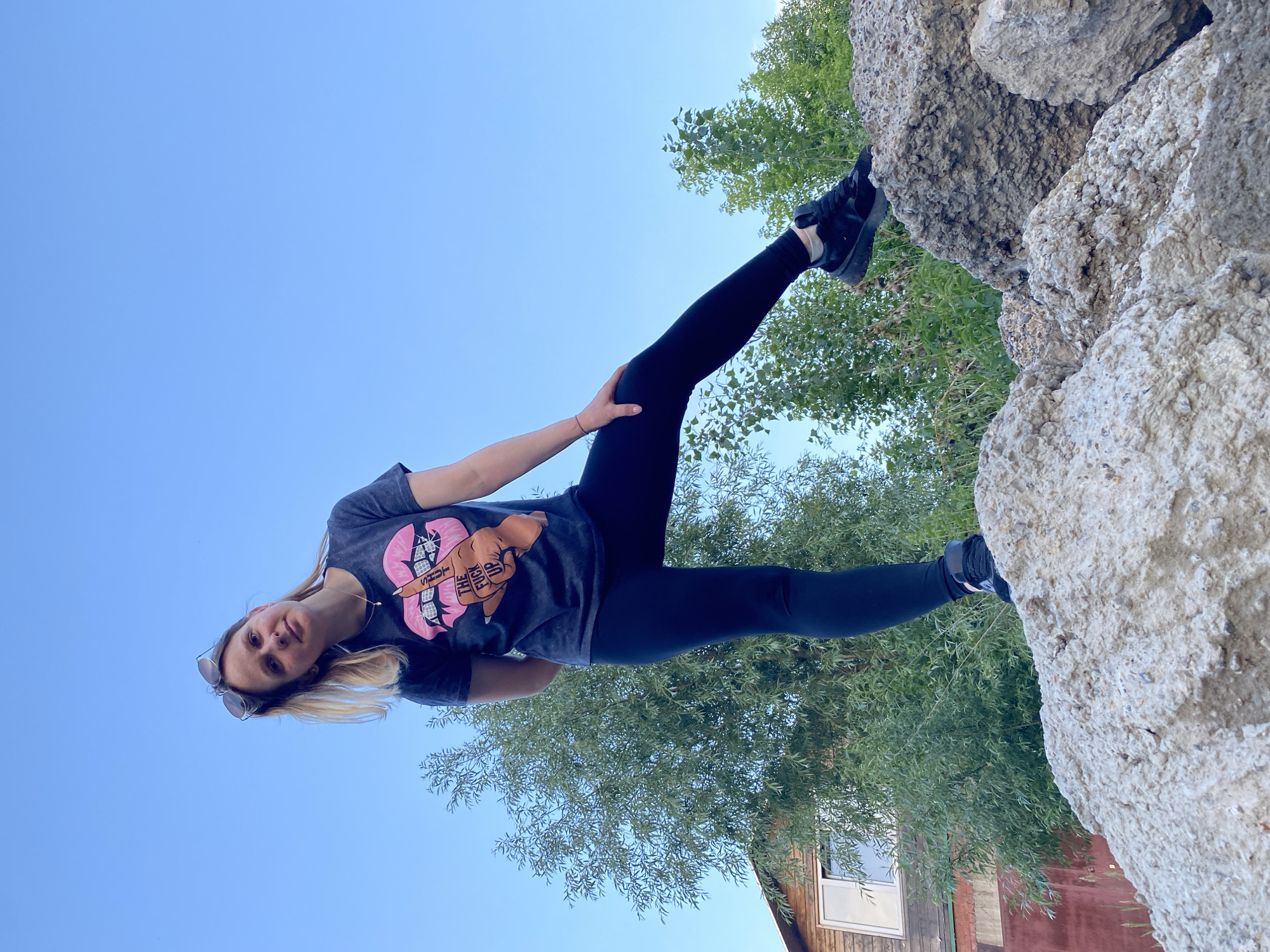A woman stands on a pile of concrete debris outdoors, with a clear blue sky in the background. She is wearing a dark t-shirt with a graphic of pink lips and a middle finger, black leggings, and black sneakers. Sunglasses are perched on her head. To her left, a large green tree obscures a portion of a wooden building. The scene is situated in a location consistent with the outskirts of Chaikovsky, Russia, possibly a construction or demolition site. The lighting suggests it is daytime, with the sun high in the sky.