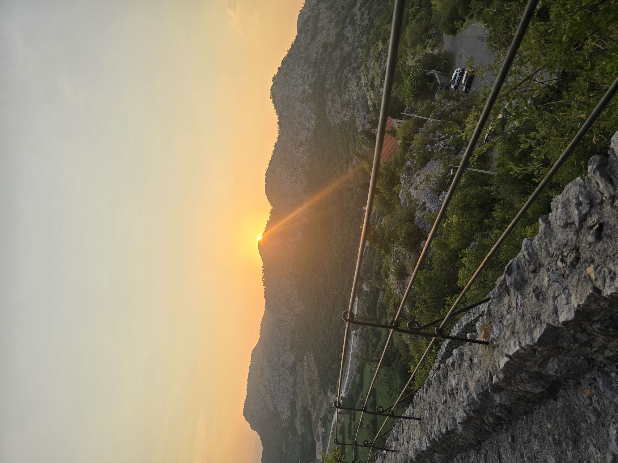 An outdoor, vertical, low-angle shot captures a sunset over the mountainous landscape of Gornja Vrbica, Montenegro. The sky transitions from a pale yellow at the bottom to a very light gray at the top. A bright orange sun, partially obscured by a mountain ridge, is low in the sky, casting golden rays across the scene. The foreground features a textured stone wall and a metal railing. Beyond the railing, lush green trees cover rolling hills and mountains. A winding road with two parked cars is visible in the middle ground, along with a few small buildings.
