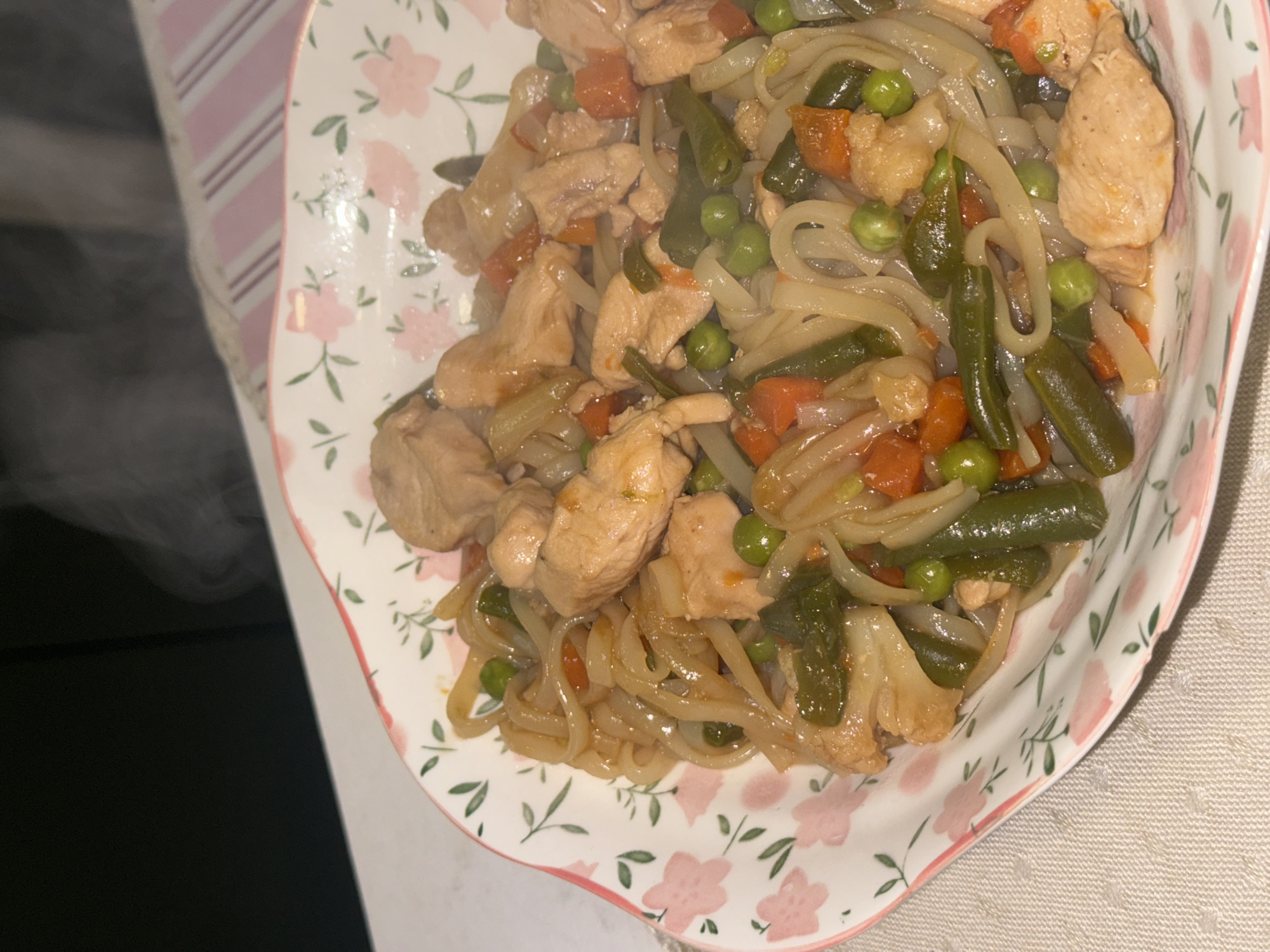 A close-up shot shows a floral patterned bowl filled with a stir-fry-like dish. The dish features noodles, cooked chicken pieces, green beans, carrots, cauliflower and peas. Steam rises from the food. The location of the photo is unknown, therefore I cannot confirm it is in Krasnoyarsk, Russia.