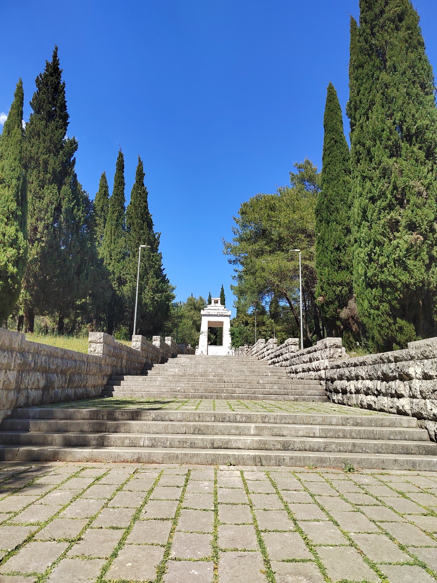 The image depicts a monumental stone stairway leading towards a white structure in a park-like setting, under a clear blue sky. Tall, slender trees line the sides of the stairway, creating a sense of enclosure and formality. The stairway itself is made of wide, shallow steps constructed from light-colored stone. The steps rise from a paved area with square stone blocks, and patches of grass grow between the stones. Low stone walls border the stairway on either side. The white structure at the top of the stairs appears to be a monument or pavilion, with a simple, open design.
Based on the architectural style and environment, the image likely depicts the Partisan Monument on Gorica Hill in Podgorica, Montenegro. The monument commemorates the Partisan soldiers who fought during World War II.