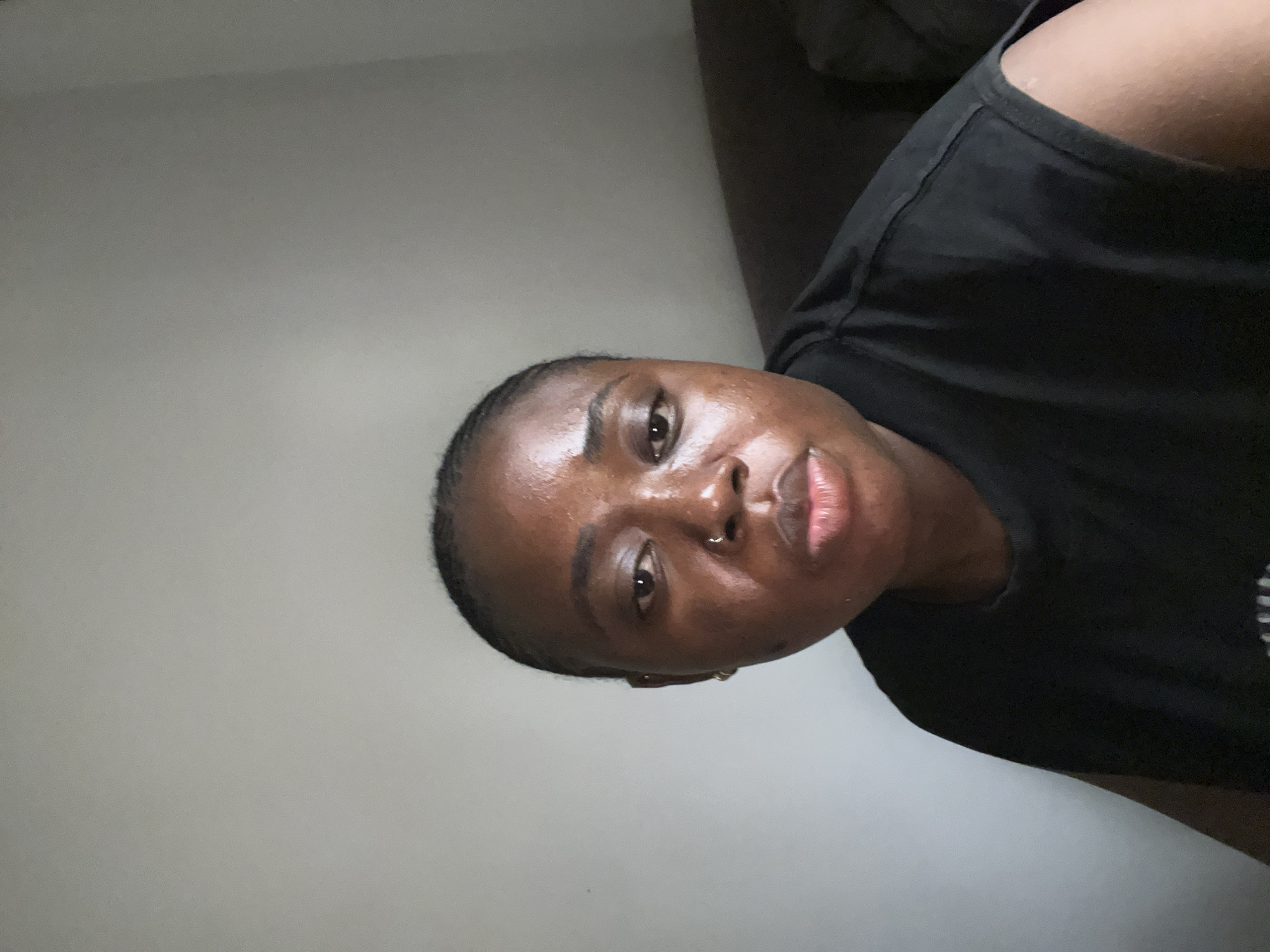 A medium shot captures a Black woman from the chest up, looking directly at the camera. She has braided hair pulled back from her face, a nose ring, and earrings. Her skin appears dewy and has a slight sheen. She is wearing a black t-shirt with wide shoulder straps. The background is a plain, light-colored wall, with a dark, textured surface, possibly a sofa, visible on the right side of the frame. The lighting is soft, with the light source appearing to be from the left, casting subtle shadows. The setting is likely an indoor environment in Abuja, Nigeria, judging by the context of the request.