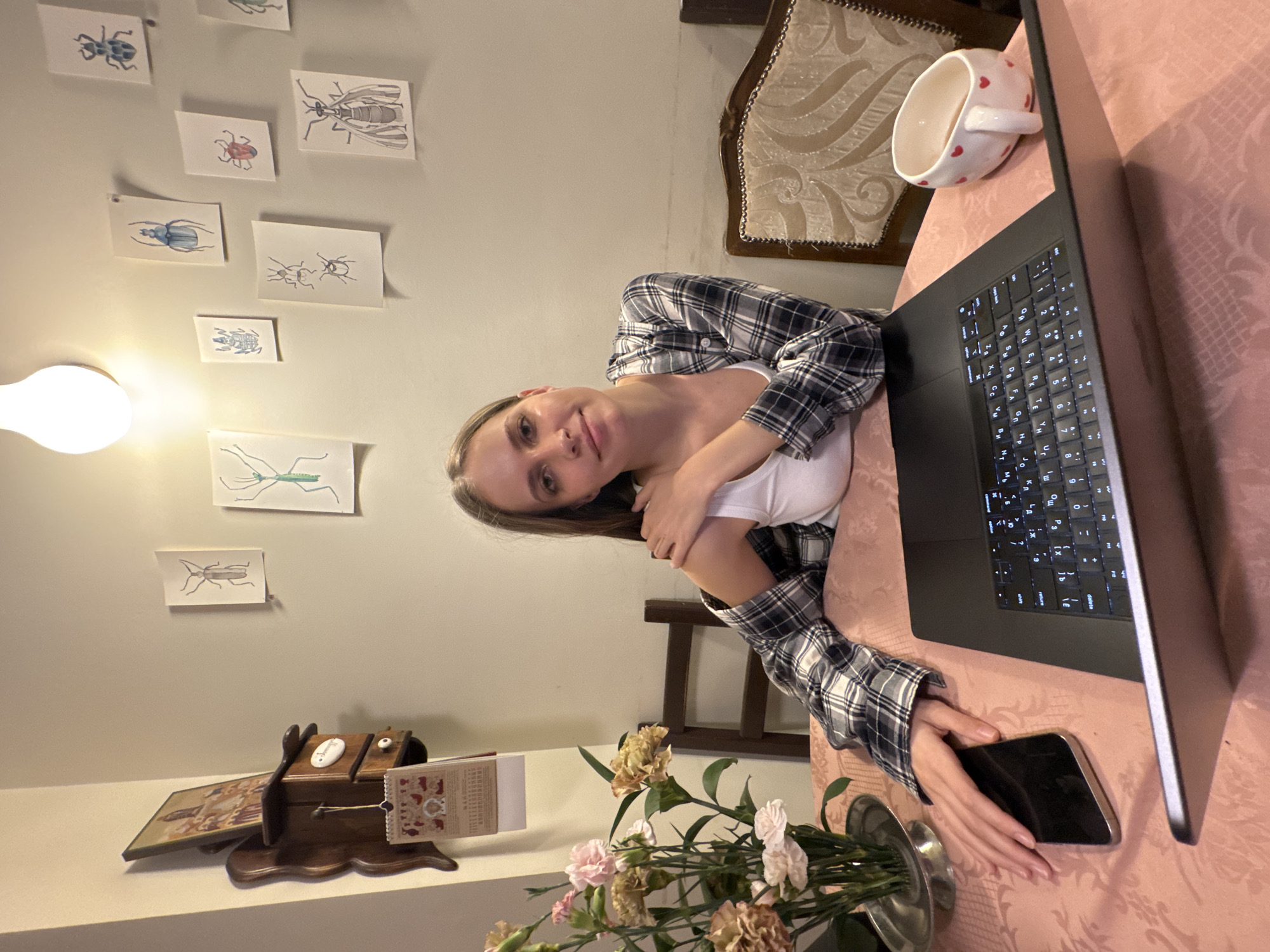 A young woman with light brown hair and a calm expression is captured in a well-lit indoor setting, likely a home, in Moscow, Russia. She is wearing a white tank top layered under a black and white plaid shirt. She is seated at a table covered with a pinkish patterned tablecloth, with her arms crossed and her head slightly tilted, looking directly at the camera with a gentle smile.
To her right, a dark-colored laptop is open on the table, its keyboard illuminated, displaying Cyrillic characters, which aligns with the given location context. A white mug adorned with red hearts sits beside the laptop. Her right hand rests on a dark-colored smartphone next to the laptop. To her left, a vase holds a bouquet of light pink and pale yellow carnations.
The background features a light-colored wall decorated with multiple small, framed illustrations of various insects, including beetles, an ant, a praying mantis, and a wasp. A prominent, modern bare lightbulb fixture provides bright illumination from the top left. Behind the woman, part of a wooden chair with an ornate upholstered back is visible. To her far left, on another surface, there is a small wooden object, possibly a decorative box or calendar holder, next to a framed picture. The artificial lighting suggests the photograph was taken in the evening or at night. The woman appears to be pausing from her activity at the laptop, posing for the camera.
