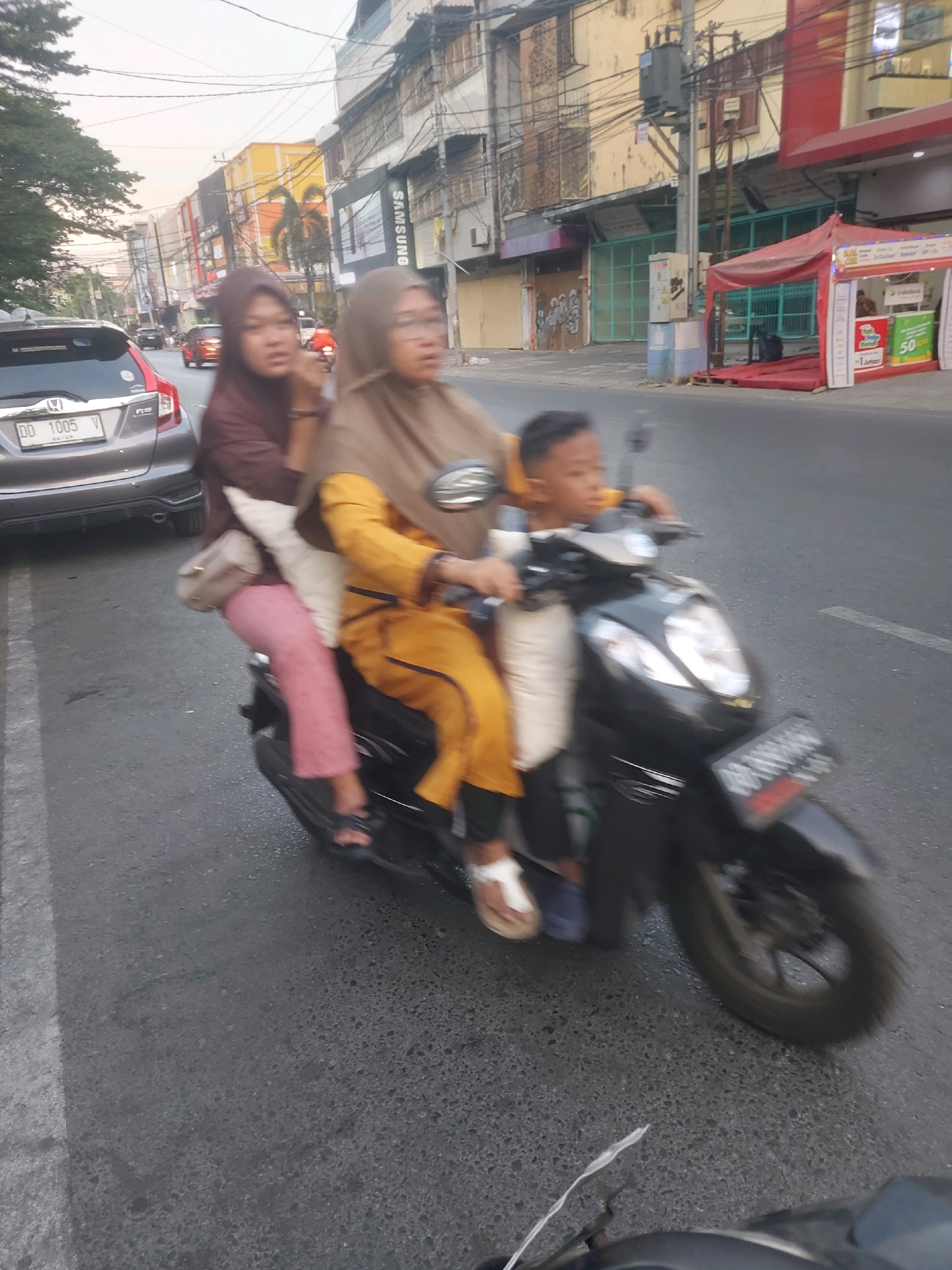 On a street in Makassar, Indonesia, three people ride a black motorcycle. A woman in a brown hijab and a yellow outfit drives, with another woman in a brown hijab and pink pants behind her, and a young boy sits between them. The motorcycle is in motion on an asphalt road with a white line. In the background, buildings with signs are visible, and power lines crisscross the sky. A silver car is parked on the left. The scene appears to be during daylight hours, with soft lighting.
