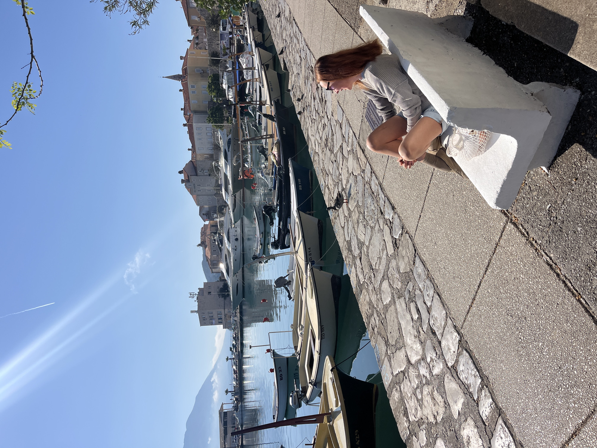 A woman with red hair is perched on a white concrete bench, her body facing the waterfront in Budva, Montenegro. She wears a grey sweater, blue denim shorts, and brown shoes, with a white string bag at her feet. Her hands are clasped together as she gazes down toward the water. The scene is bright and sunny, with clear blue skies reflecting in the water. A long line of boats of various sizes are docked along the stone-lined edge of the harbor. The building structures of the city are visible across the water, featuring stone or stucco exteriors and red-tiled roofs. Mountains form a backdrop to the city in the distance. A stone-paved walkway runs alongside the harbor, adjacent to the bench. The orientation of the image is slightly rotated counterclockwise.