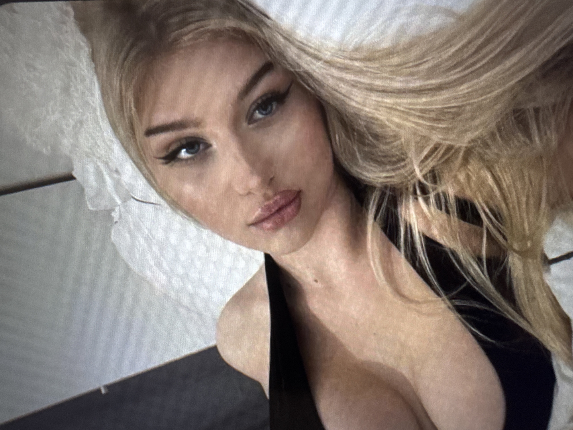 A close-up portrait depicts a young woman with long, flowing blonde hair cascading around her. She has fair skin, striking blue eyes, and prominent dark winged eyeliner, complemented by well-defined eyebrows and what appear to be long eyelashes. Her lips are full and glossy, wearing a light pink or nude shade, and are slightly parted.
She is dressed in a black garment, possibly a top or dress, with a deep neckline, revealing her décolletage. Her head is tilted, and she gazes directly at the viewer. A soft, white object, likely a pillow, is visible behind her head on the left side of the frame, suggesting an indoor setting. The overall pose is relaxed and intimate, indicative of a selfie or a close-up photograph taken from a slightly elevated angle. There is no visible text in the image.