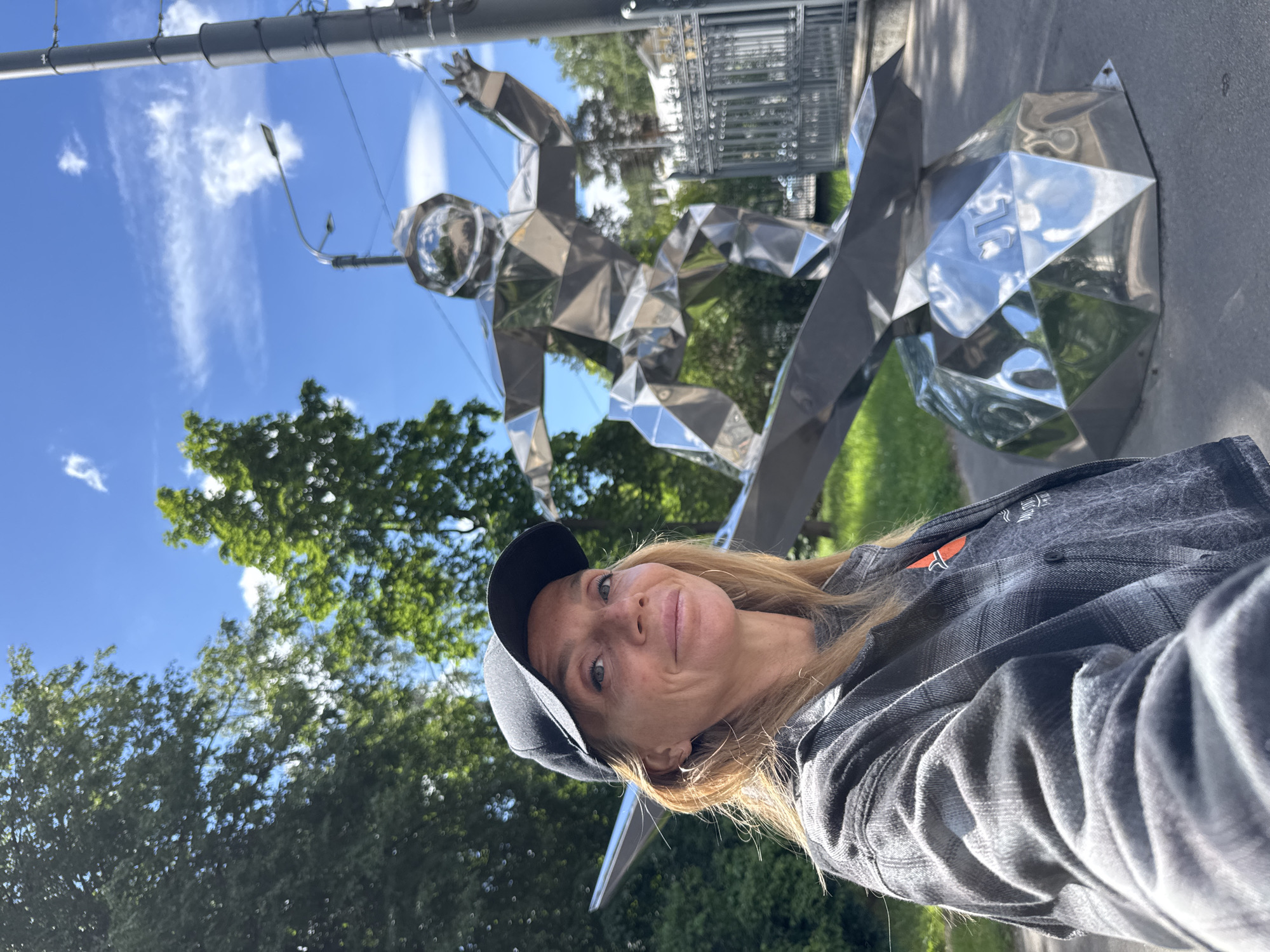 The media file captures a woman in a selfie, posing outdoors. She is wearing a dark baseball cap and a plaid shirt. Her hair is blonde, and she is smiling at the camera. Behind her, a large, abstract, metallic sculpture resembling an astronaut is prominently displayed. The sculpture is made of many geometric facets, reflecting the surrounding environment. The scene is set under a bright blue sky with scattered white clouds. Lush green trees are visible in the background, suggesting a park or outdoor public space. A lamppost and other street furniture are also present. The lighting indicates it is daytime, likely late morning or afternoon, with sunny weather. The overall mood appears cheerful and relaxed. There is no visible text in the media file. The location context provided is Sinyavino, Russia, which might suggest this is a public art installation in that area.