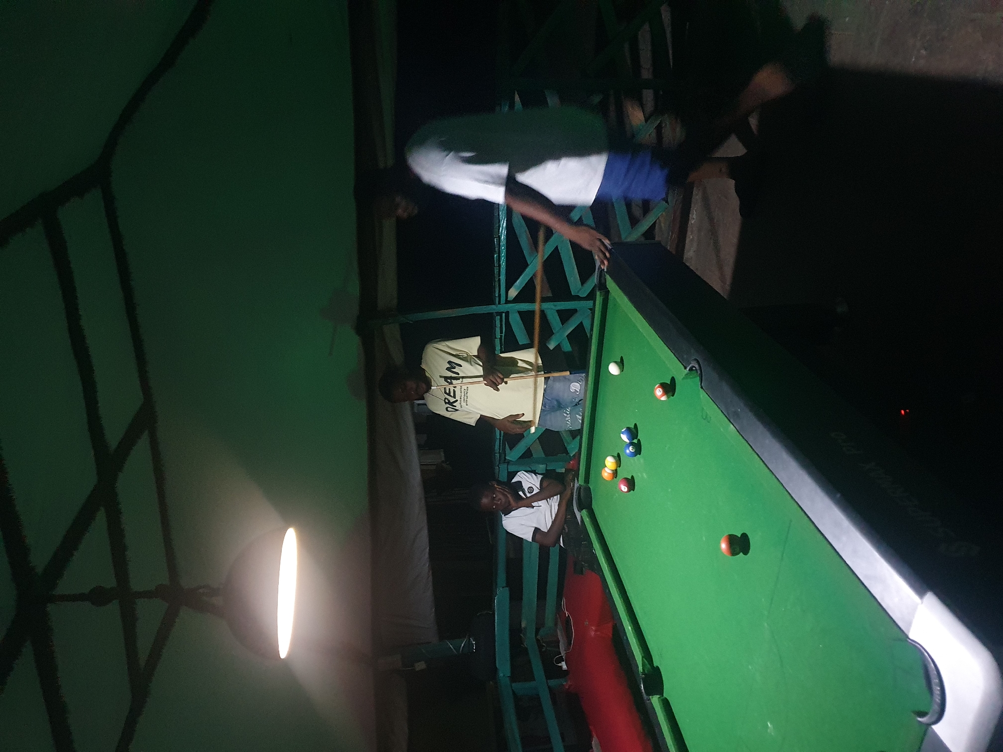 A group of three young men are playing pool at night, outdoors, under a covered structure. The scene is illuminated by a single overhead light. A green pool table with several colored balls is the central object. One man, wearing a white t-shirt, stands to the right of the table, his hand near the edge of the table. Another man, in a yellow t-shirt with "DREAM" printed on it, stands behind the table, holding a pool cue. A third, younger man, wearing a white polo shirt, sits on a bench to the left of the table, looking at the table with his hand on his chin. The background shows a dark, outdoor environment with a wooden railing and a canvas covering overhead. The ground appears to be concrete. The overall impression is of a casual, nighttime gathering.