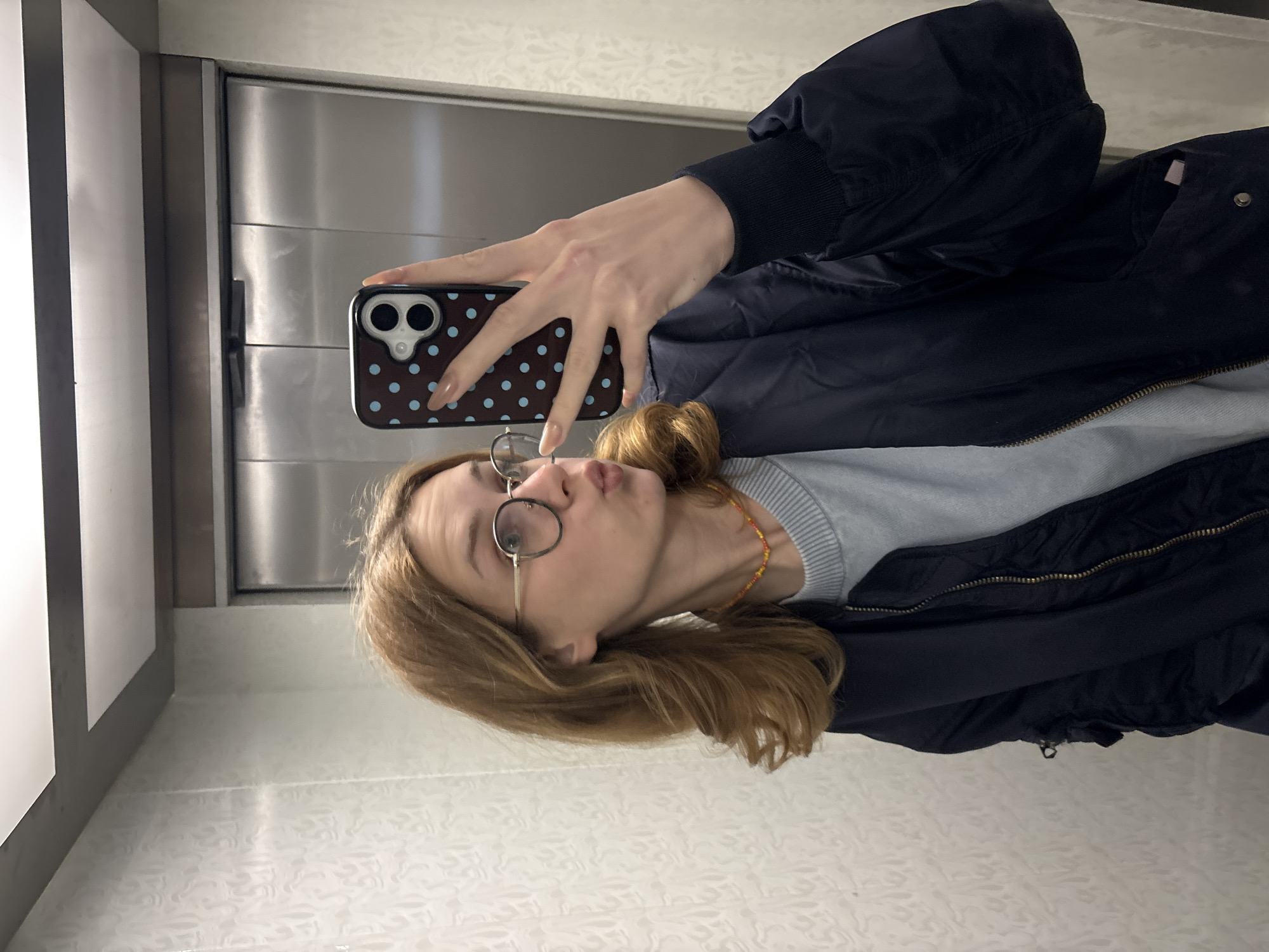 The image is a selfie depicting a young person, likely female, seen from the chest up, captured in what appears to be a mirror or reflective surface. The person has shoulder-length light brown hair and wears round wire-frame glasses. She is making a playful "kissing" or puckered face towards the camera. Her attire includes a light blue/grey ribbed long-sleeved top underneath a dark blue/black bomber jacket. A colorful beaded necklace, featuring red, yellow, and orange beads, is visible around her neck.
She holds a smartphone to take the picture, with her hand partially obscuring her face. The phone has a distinctive dark brown case covered in light blue polka dots, and its camera lenses are clearly visible. Her fingernails have a light, natural-toned polish.
The background suggests an indoor setting, likely within an elevator or a small room in Kragujevac, Serbia. To the left, a set of metallic doors or wall panels are visible, indicating an industrial or modern interior. The right side and bottom of the frame reveal a light-colored wall or floor with a subtle, intricate pattern, adding a textural element to the otherwise simple environment. No text is visible in the image.