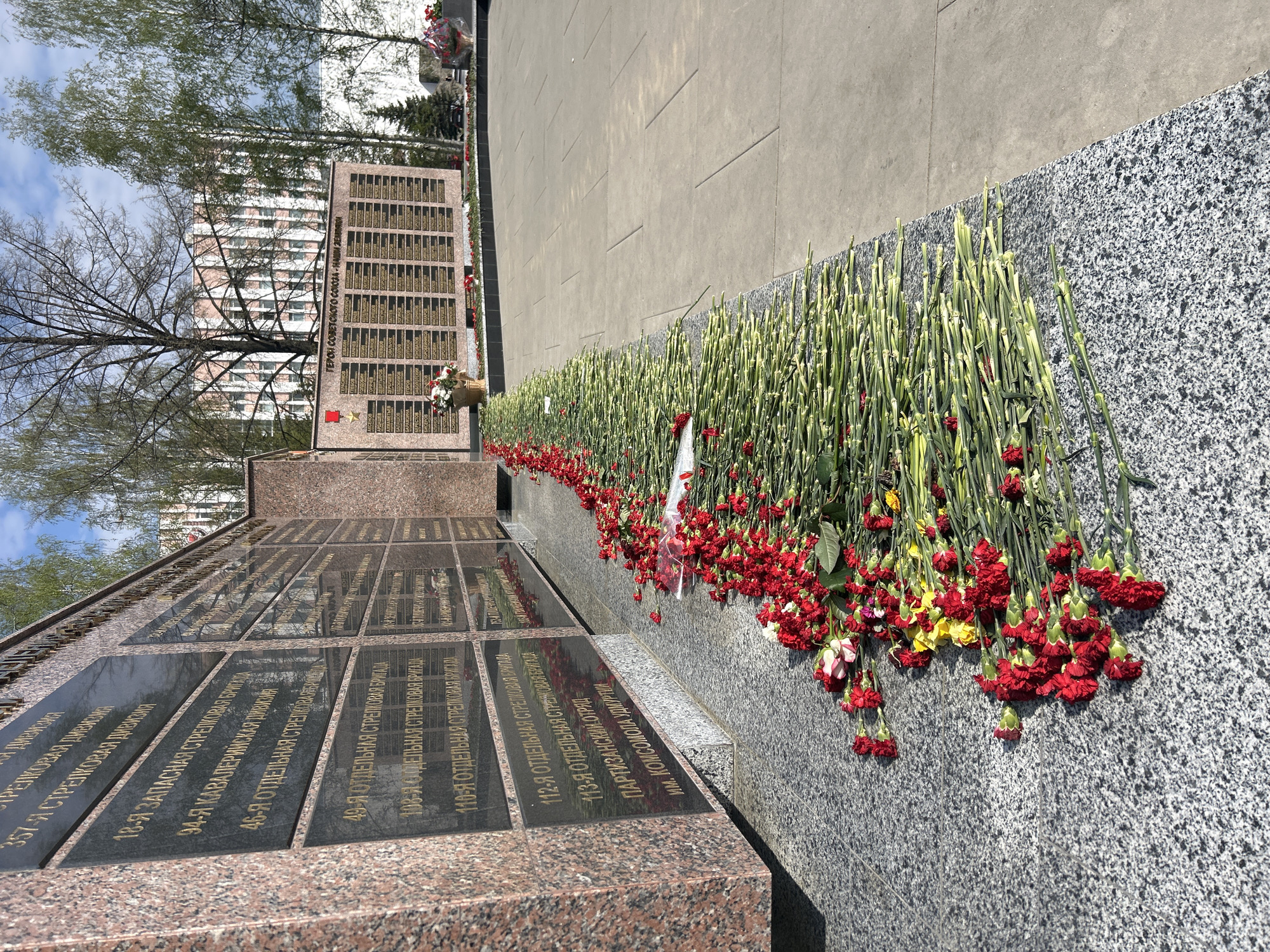 The image shows a memorial in Izhevsk, Russia. The memorial consists of multiple black plaques with gold lettering mounted on granite structures. The plaques appear to list military units. At the base of the memorial, along a gray stone surface, are numerous red and yellow carnations laid in remembrance. In the background, there are trees and a building. The weather appears to be sunny with some clouds in the sky.