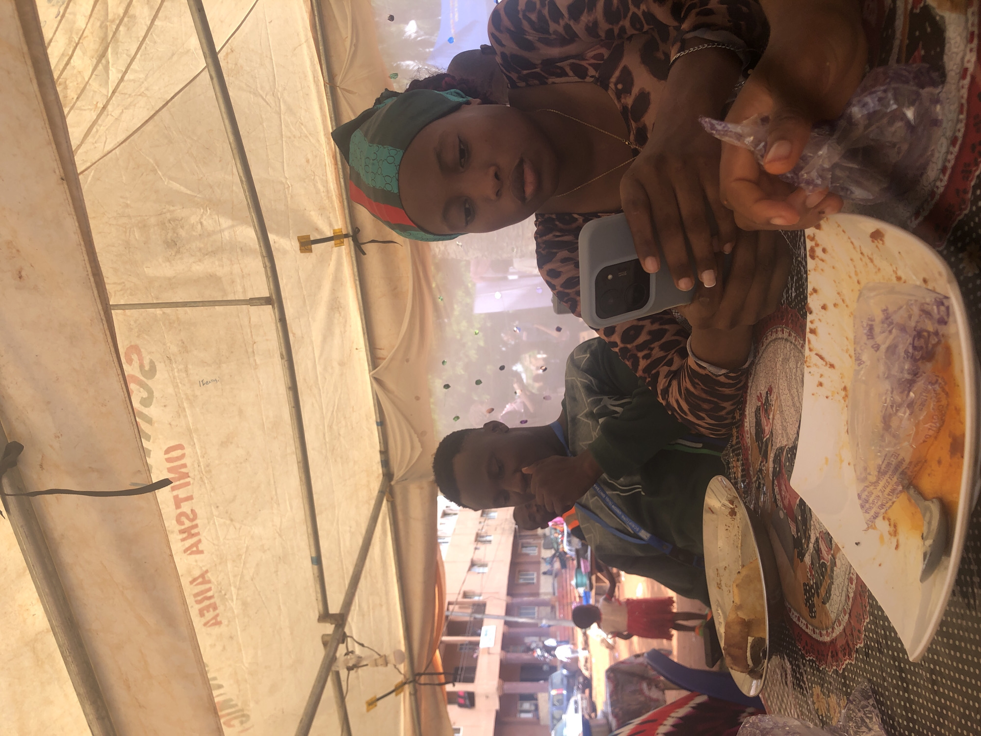 Two individuals are seated at a table, likely under a tent, in Nteje, Nigeria. The woman, wearing a leopard print top and a headwrap, holds a mobile phone and a plastic bag. A man sits across from her, wearing a green top and a lanyard. The table has plates of food, some partially consumed. The background reveals a street scene with buildings and other people.