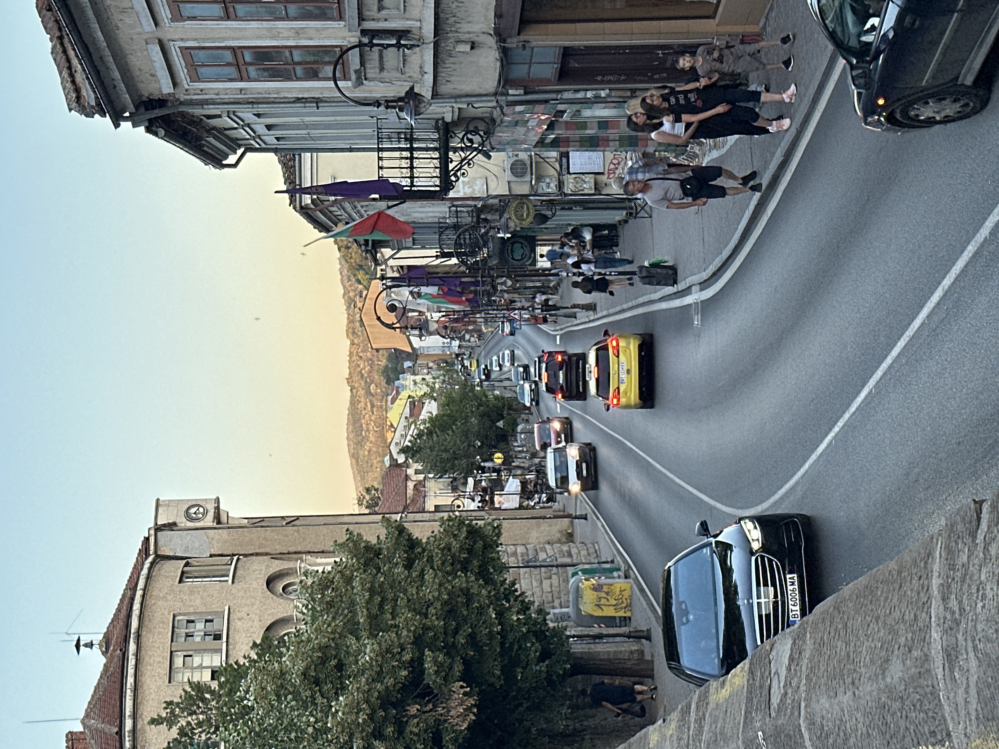 This image captures a street scene in Veliko Tarnovo, Bulgaria, likely in the old town area, characterized by its hilly terrain and historic architecture. The perspective is from an elevated vantage point, looking down a sloped street filled with traffic and pedestrians.
In the foreground, a dark sedan with license plate "BT 6006 MA" is visible on the right side of the road, partially obscured by a stone wall on the left. The road itself is asphalt with white lane markings and appears to be a two-way street with multiple lanes.
Further up the street, various vehicles, including a yellow sedan, a white van, and several other cars and SUVs, are moving or parked. The street is lined with buildings on both sides. On the left, a large, multi-story building with a curved facade and a prominent clock tower is visible. Adjacent to it are trees and street furniture, including trash bins.
The right side of the street features older buildings with balconies, ornate street lamps, and shopfronts. Several Bulgarian flags are displayed on the buildings, indicating a national or local celebration. Pedestrians are walking on the sidewalks, some carrying bags or luggage, suggesting a busy commercial or tourist area.
In the background, the street continues uphill, revealing more buildings and distant hills bathed in the soft light of what appears to be late afternoon or early evening. The sky is clear with a gradient of blue and a touch of orange from the setting sun. The overall atmosphere suggests a lively urban environment with a blend of historical charm and modern activity.