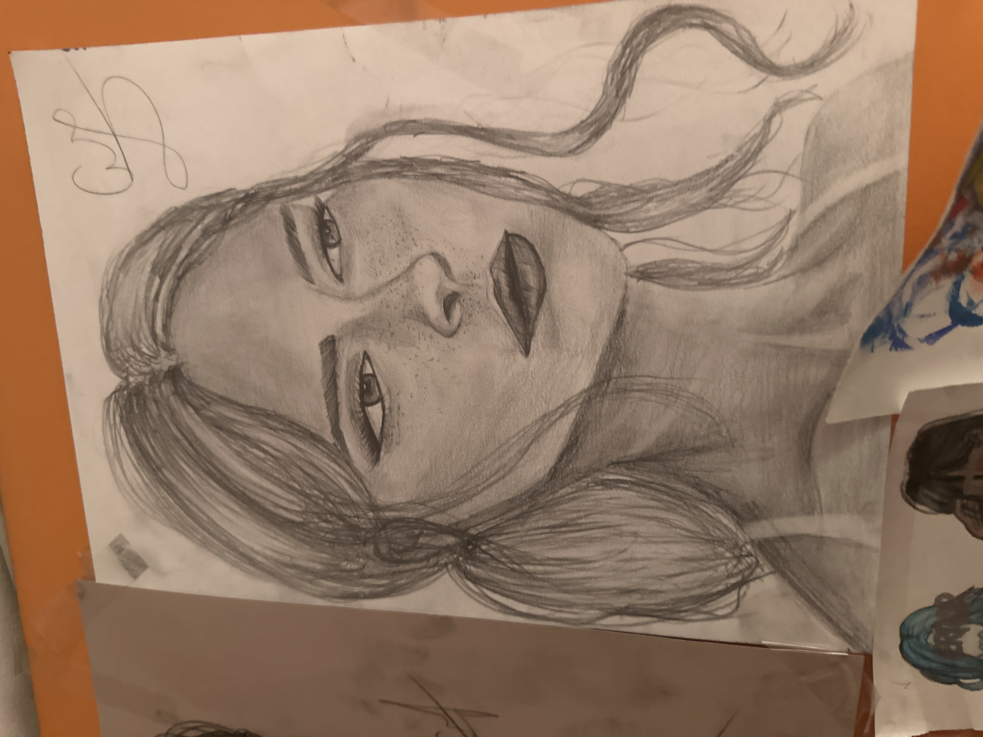 The image is a close-up, eye-level, medium shot of a pencil drawing of a woman's face. The drawing is on white paper, and the woman is depicted from the chest up, looking to her right. She has dark hair styled in a middle part with two pigtails and loose strands framing her face. She has noticeable freckles scattered across her nose and cheeks, and her lips are full and painted with dark lipstick. Her eyes are defined with eyeliner, and her eyebrows are arched. There is a stylized signature in the upper right corner of the drawing. The drawing is taped to a surface, partially obscuring another drawing to its left. Below the main drawing, the edges of two other drawings are visible, one with blue hair and another with dark hair. The background is a plain, light surface, with an orange surface visible along the top and bottom edges of the frame. The image does not contain any elements that would allow for identification of a specific location in Plovdiv, Bulgaria.